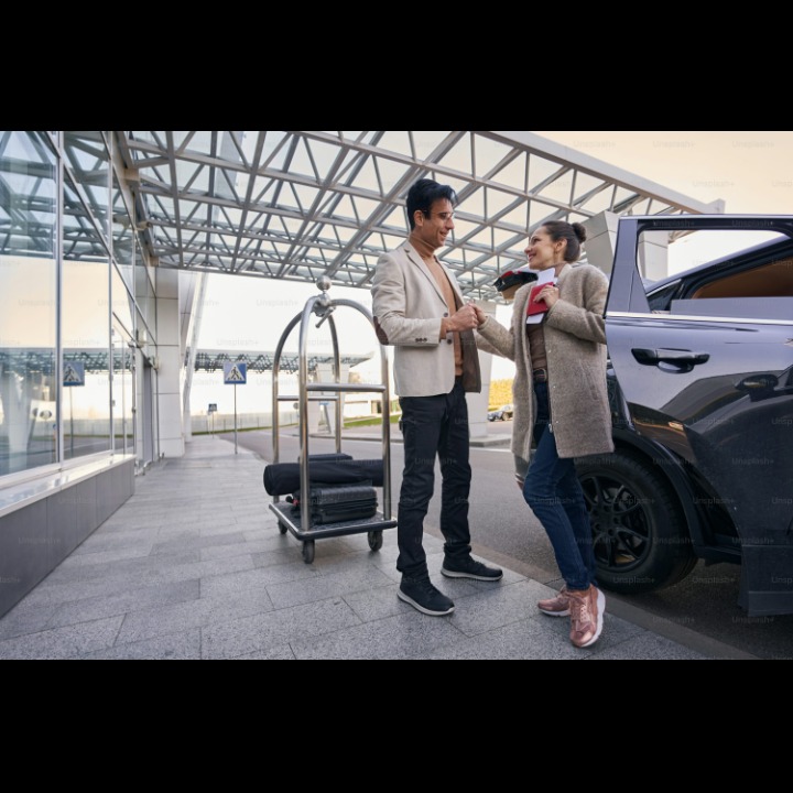 Elegancki mężczyzna w beżowej marynarce wita kobietę z paszportem przy czarnym samochodzie. W tle wózek bagażowy i przeszklony budynek lotniska.