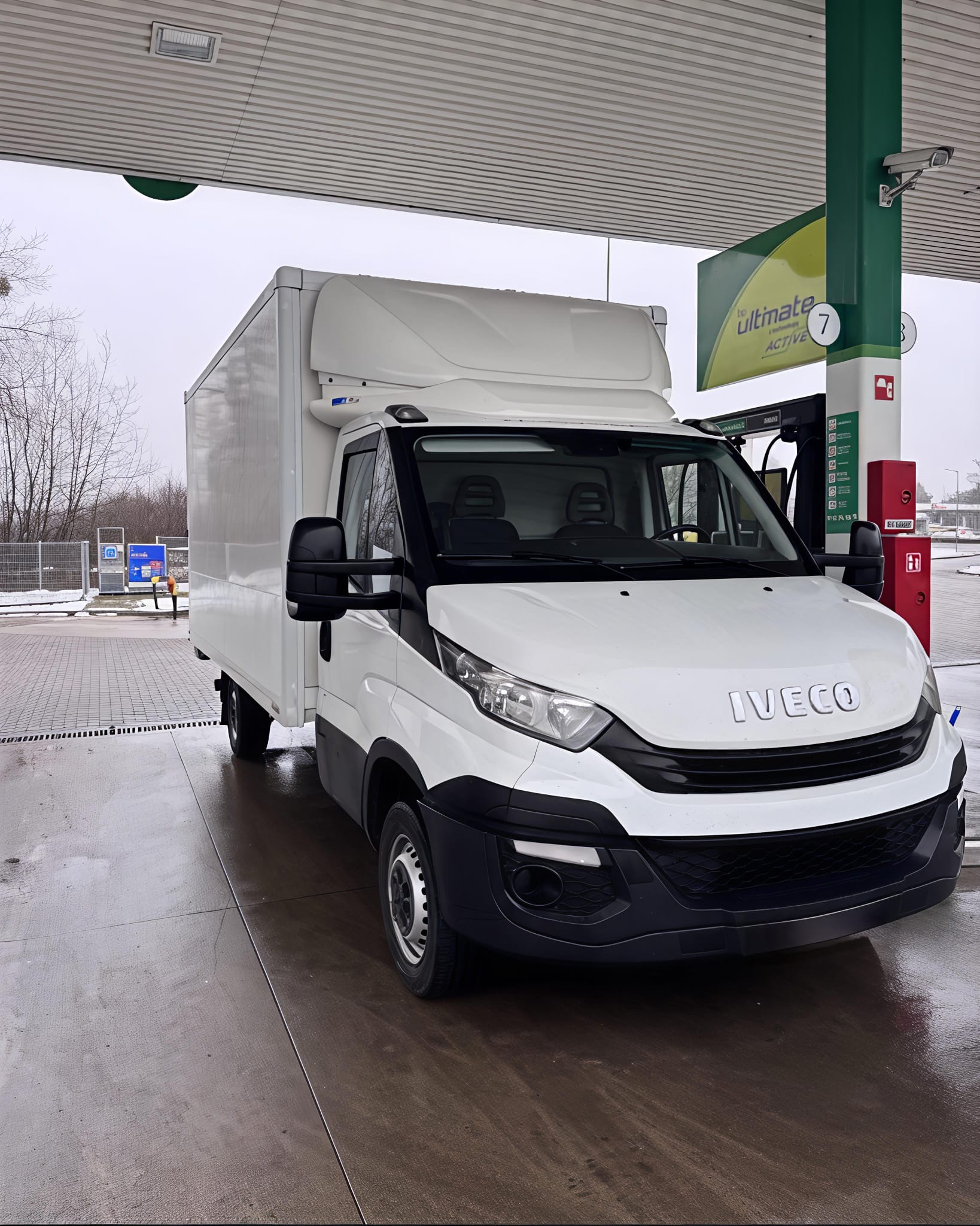 Biały samochód dostawczy Iveco z zabudową kontenerową na stacji benzynowej, widok z przodu, pochmurny dzień. Mokra nawierzchnia.