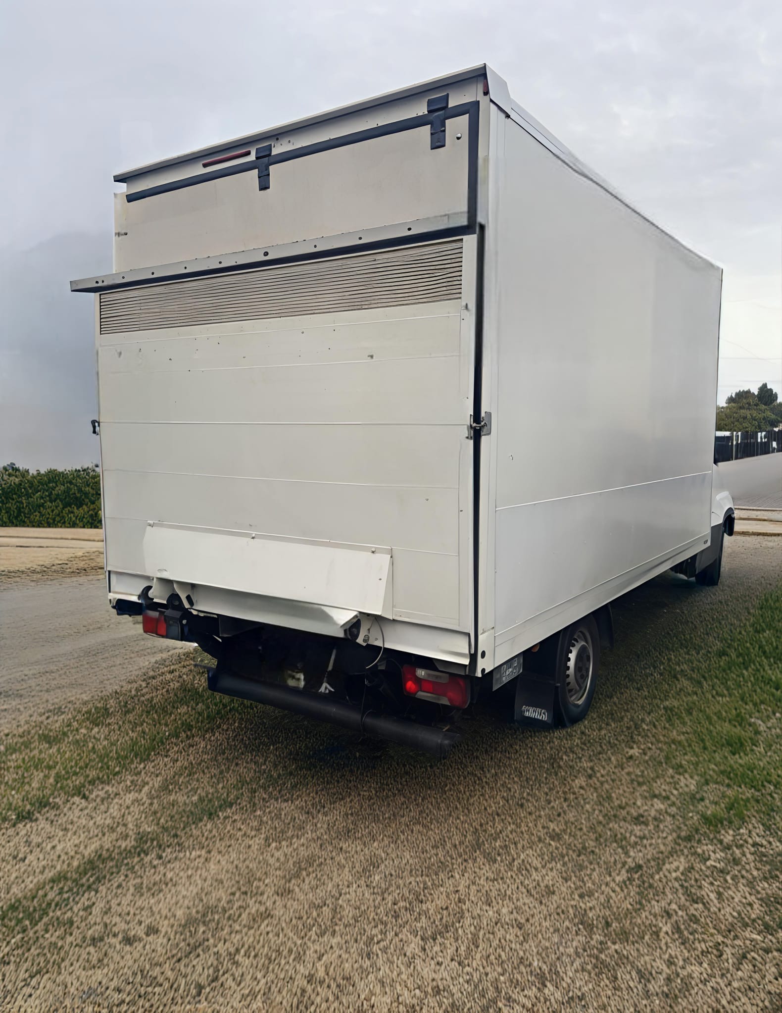 Biały samochód dostawczy z otwartymi drzwiami z tyłu, zaparkowany na trawiastym terenie. Widok z tyłu pojazdu, z widocznymi detalami karoserii i oświetlenia.