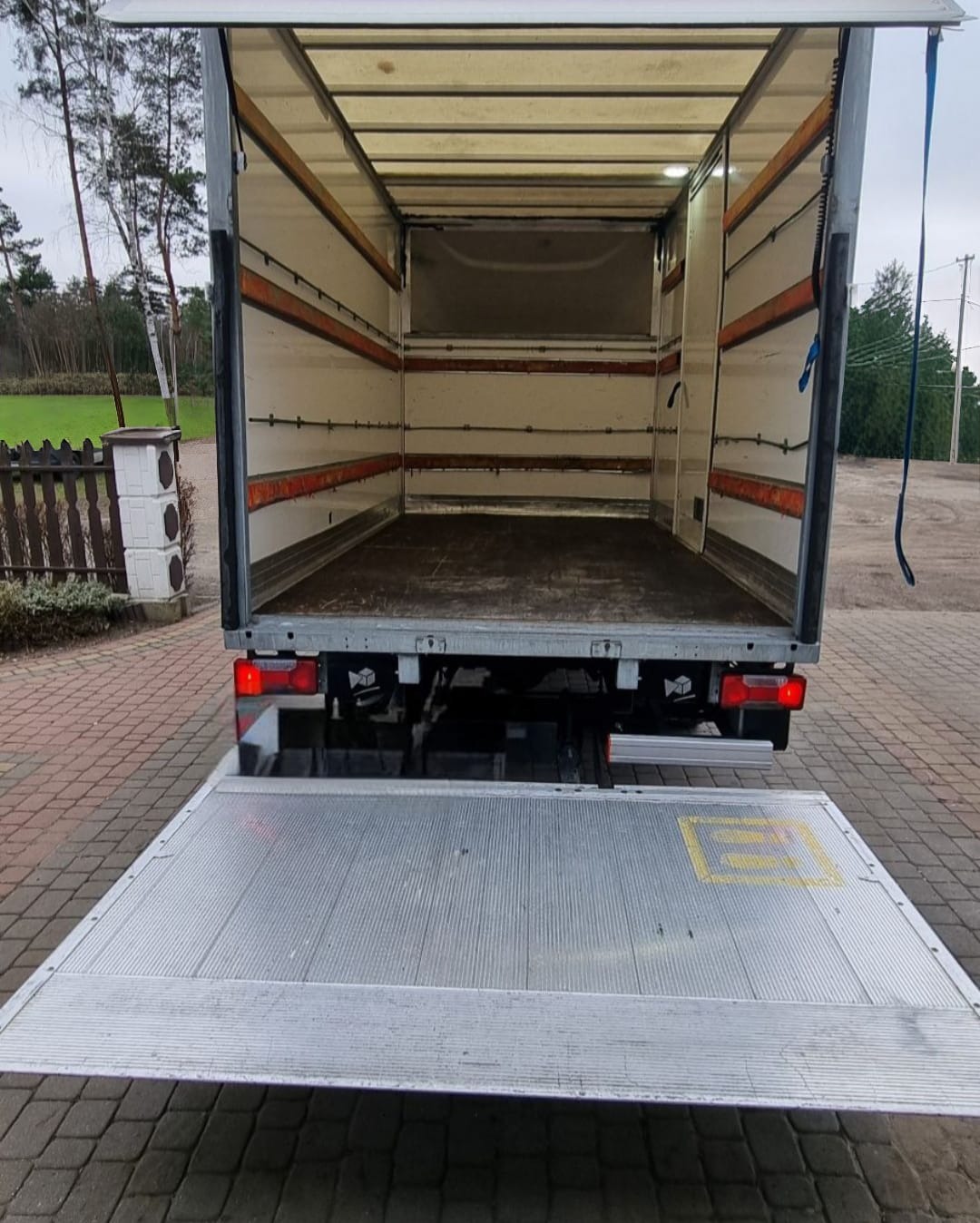 Wnętrze paki busa transportowego z otwartą rampą załadunkową, widok z tyłu na zewnątrz, ceglana kostka brukowa na podłożu.