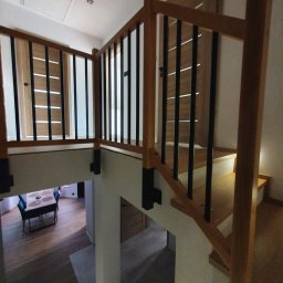 Rodek Kamil - Nowoczesne schody z drewnianą balustradą z czarnymi prętami, widok z góry na dół, w tle fragment salonu z drewnianą podłogą i stołem.