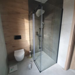 Rodek Kamil - Nowoczesna kabina prysznicowa z czarnymi profilami i deszczownicą, drewnopodobne płytki na ścianie, podwieszana toaleta. Wykończenie łazienki w szarościach i drewnie.