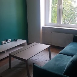 Rodek Kamil - Salon po odnowieniu: zielona ściana, jasne meble, drewniana podłoga i okno z widokiem na zieleń. Nowoczesny design wnętrza z akcentem na kolor i naturalne światło.