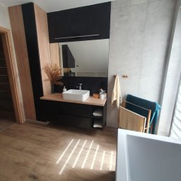 Rodek Kamil - Nowoczesna łazienka z drewnopodobną podłogą, umywalką na szafce z czarnymi frontami i szarą ścianą z dużych płytek. W tle widoczna wanna i grzejnik drabinkowy.