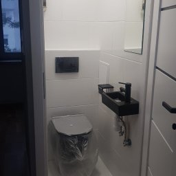 Rodek Kamil - Wąska łazienka z białą glazurą, czarną armaturą i wiszącą umywalką. Toaleta zabezpieczona folią ochronną. Widoczny licznik gazu.