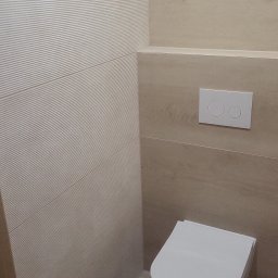 Rodek Kamil - Nowoczesna toaleta podwieszana w jasnej łazience, z beżowymi płytkami imitującymi drewno i strukturalną ścianą. Minimalistyczny design i czysta estetyka.