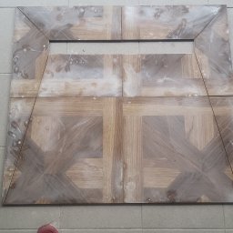 Rodek Kamil - Ułożone płytki imitujące drewno w geometryczny wzór z widocznymi śladami zaprawy, na podłodze z szarych płytek. Widoczne buty osoby fotografującej.