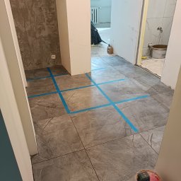 Rodek Kamil - Szare płytki podłogowe z niebieską taśmą wyznaczającą wzór układania, widok na remontowane pomieszczenia i narzędzia glazurnicze, etap prac wykończeniowych.