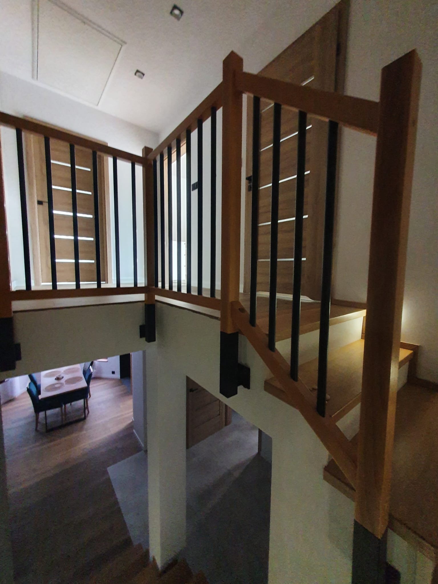 Nowoczesne schody z drewnianą balustradą z czarnymi prętami, widok z góry na dół, w tle fragment salonu z drewnianą podłogą i stołem.