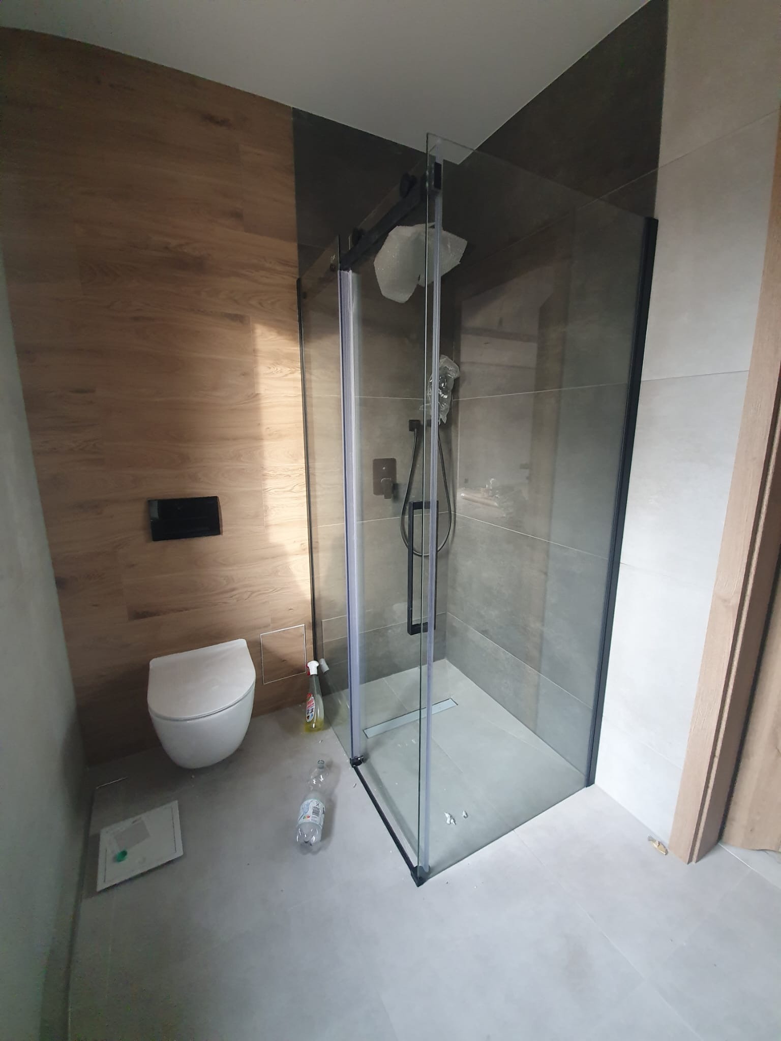 Nowoczesna kabina prysznicowa z czarnymi profilami i deszczownicą, drewnopodobne płytki na ścianie, podwieszana toaleta. Wykończenie łazienki w szarościach i drewnie.