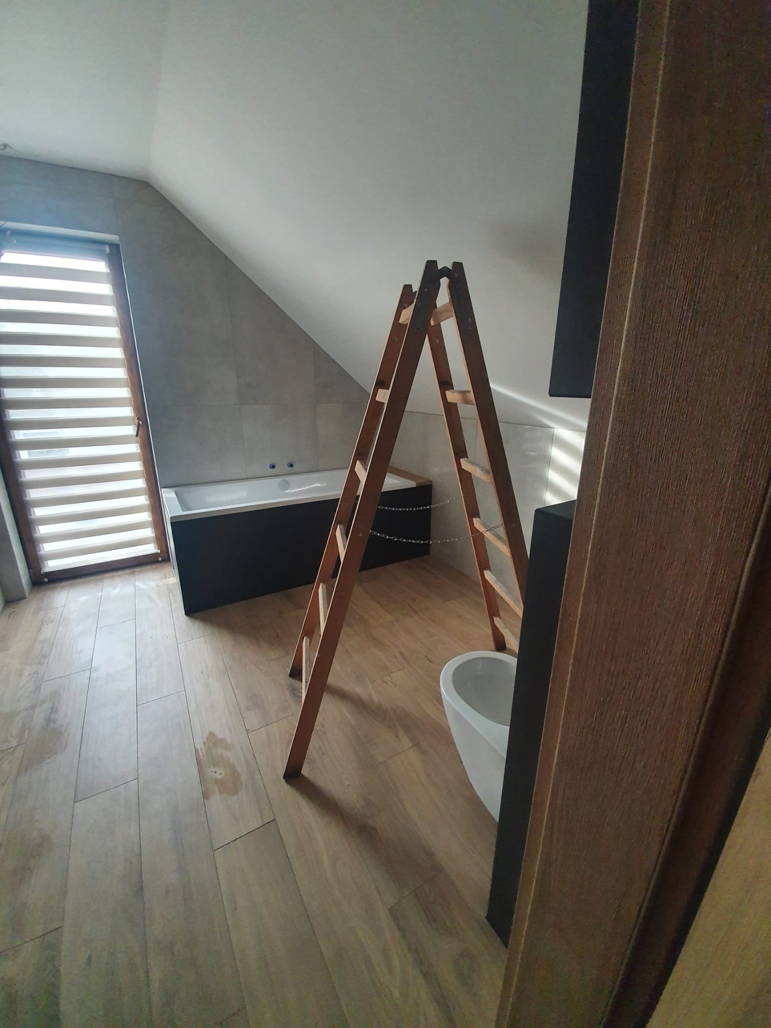 Ukończona łazienka na poddaszu z drewnianą podłogą, wanną i oknem z żaluzjami. Drabina oparta o ścianę sugeruje prace wykończeniowe.