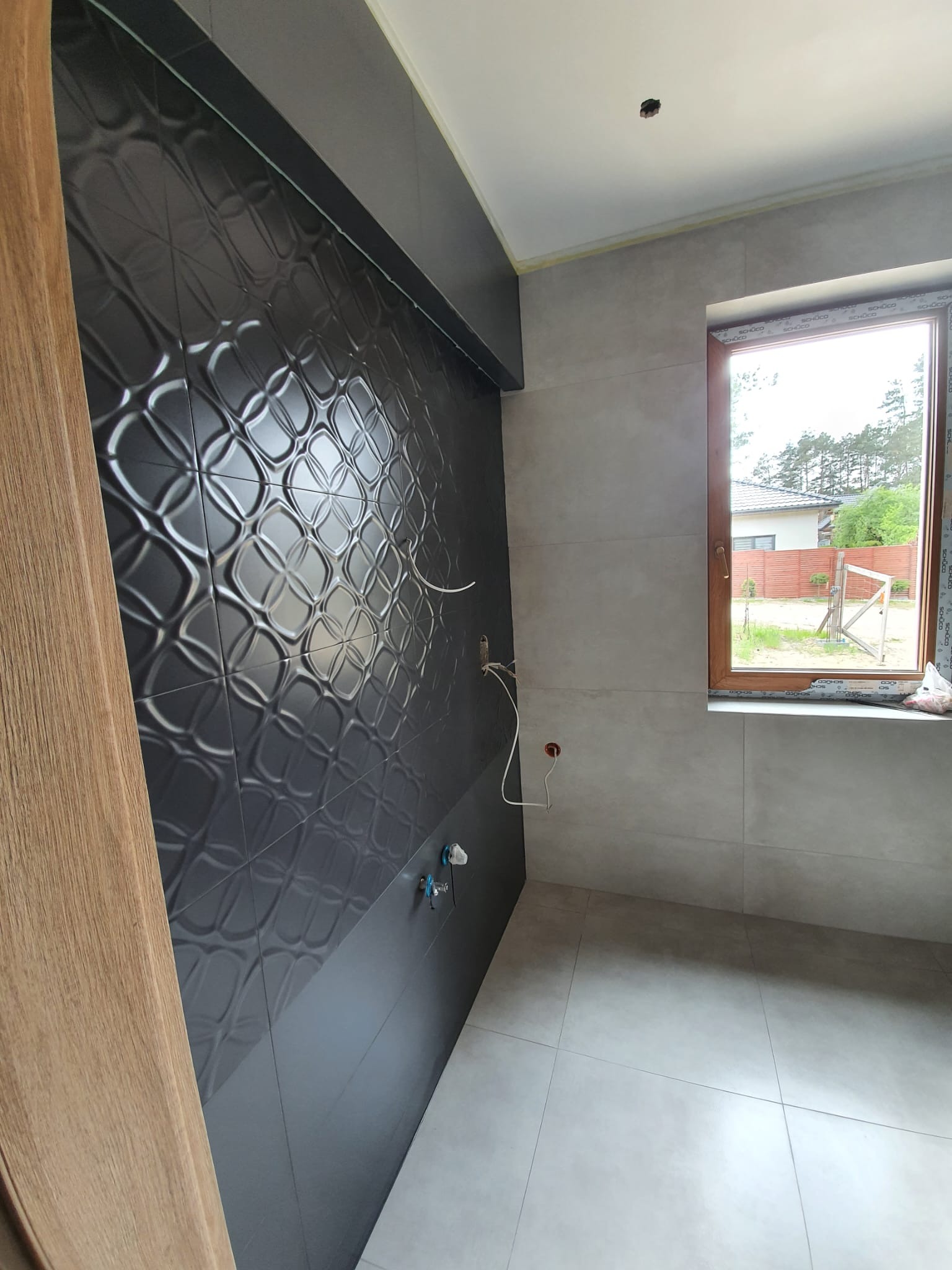 Częściowo wykończona łazienka z dekoracyjnymi, ciemnymi płytkami na ścianie i jasnymi płytkami podłogowymi. Widoczne instalacje i okno z widokiem na zewnątrz.