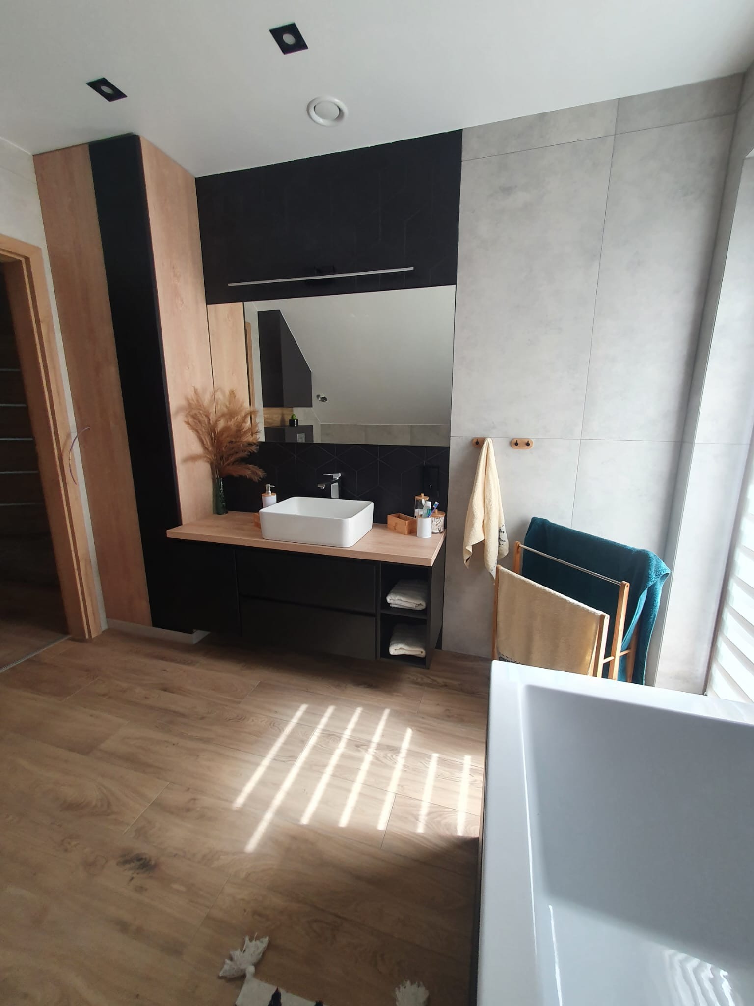 Nowoczesna łazienka z drewnopodobną podłogą, umywalką na szafce z czarnymi frontami i szarą ścianą z dużych płytek. W tle widoczna wanna i grzejnik drabinkowy.