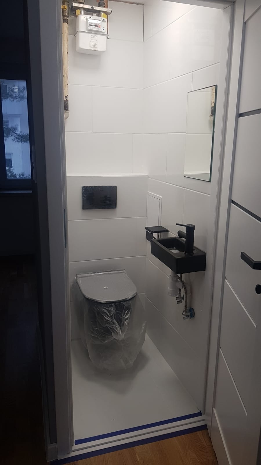 Wąska łazienka z białą glazurą, czarną armaturą i wiszącą umywalką. Toaleta zabezpieczona folią ochronną. Widoczny licznik gazu.