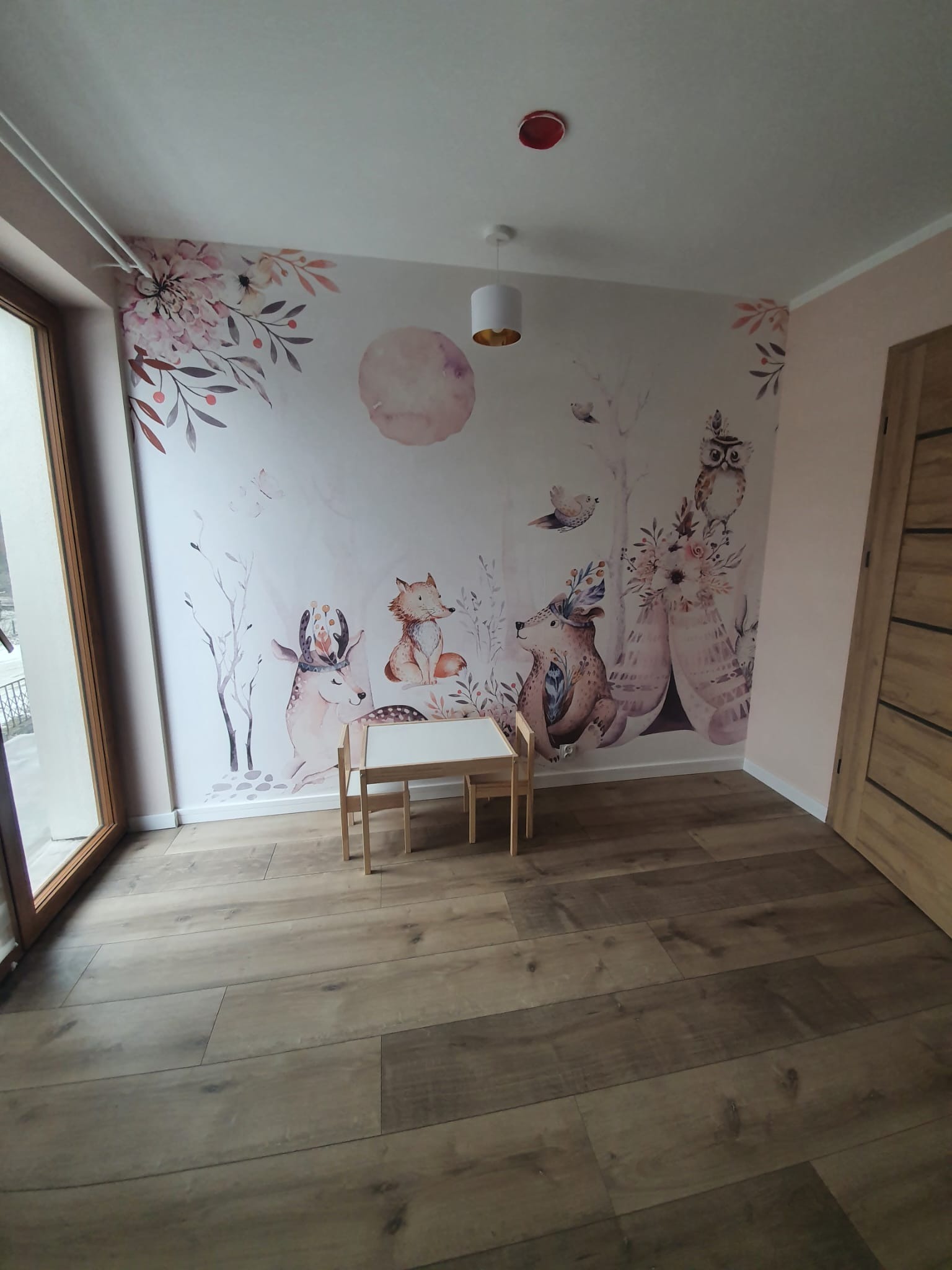 Pokój dziecięcy z fototapetą w leśne zwierzęta, drewnianą podłogą i małym stolikiem. Dekoracja ścian w pastelowych barwach, jasne wnętrze.