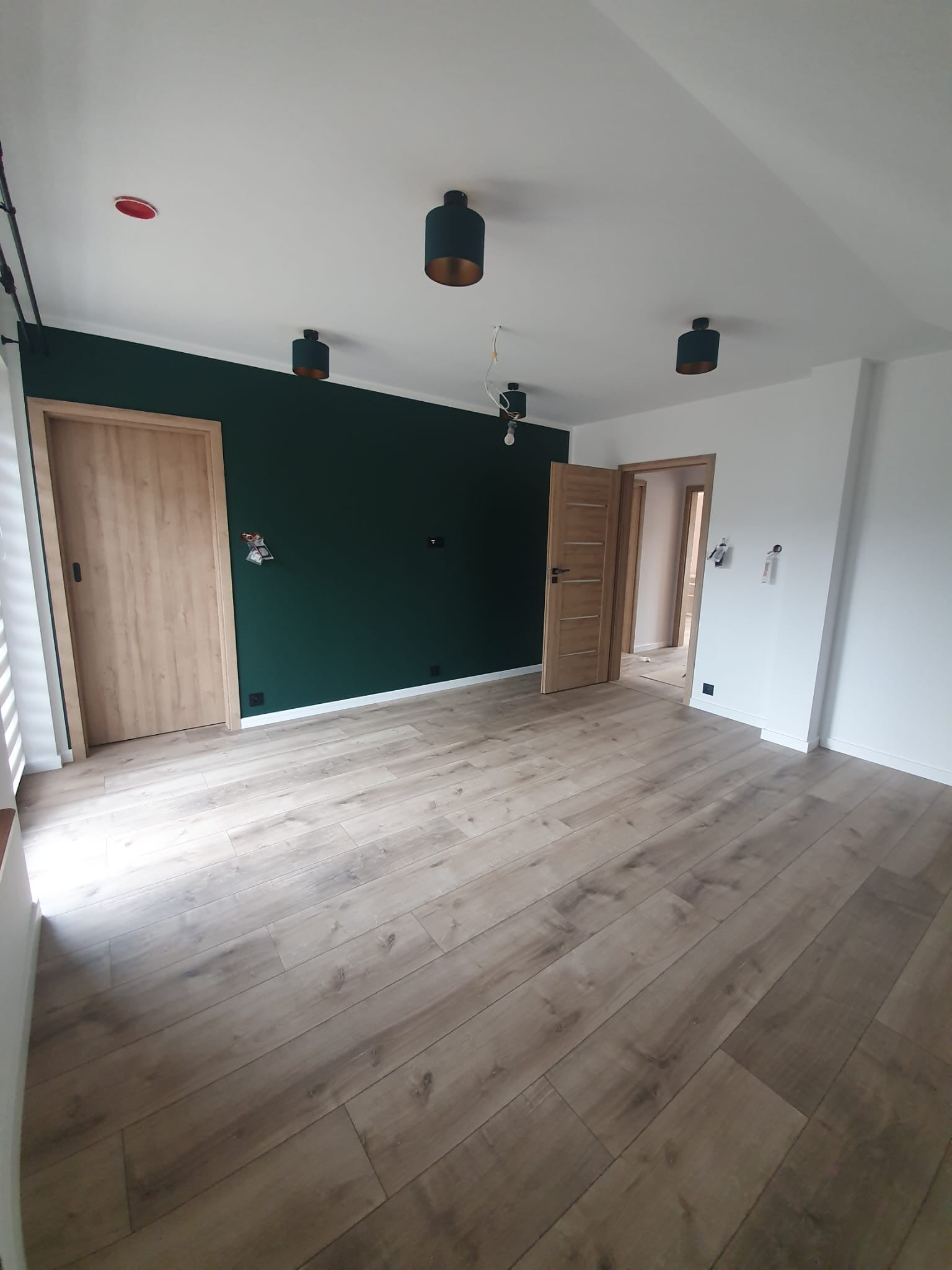 Jasne panele podłogowe w przestronnym wnętrzu z dwiema drewnianymi ościeżnicami i ścianą w kolorze butelkowej zieleni. Widoczne lampy sufitowe.