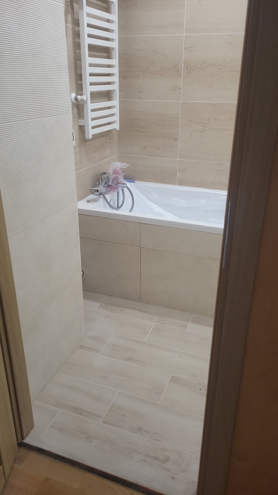 Jasna łazienka po remoncie: beżowe płytki imitujące drewno na ścianach i podłodze, biały grzejnik drabinkowy, wanna z obudową, kabina prysznicowa w folii ochronnej.