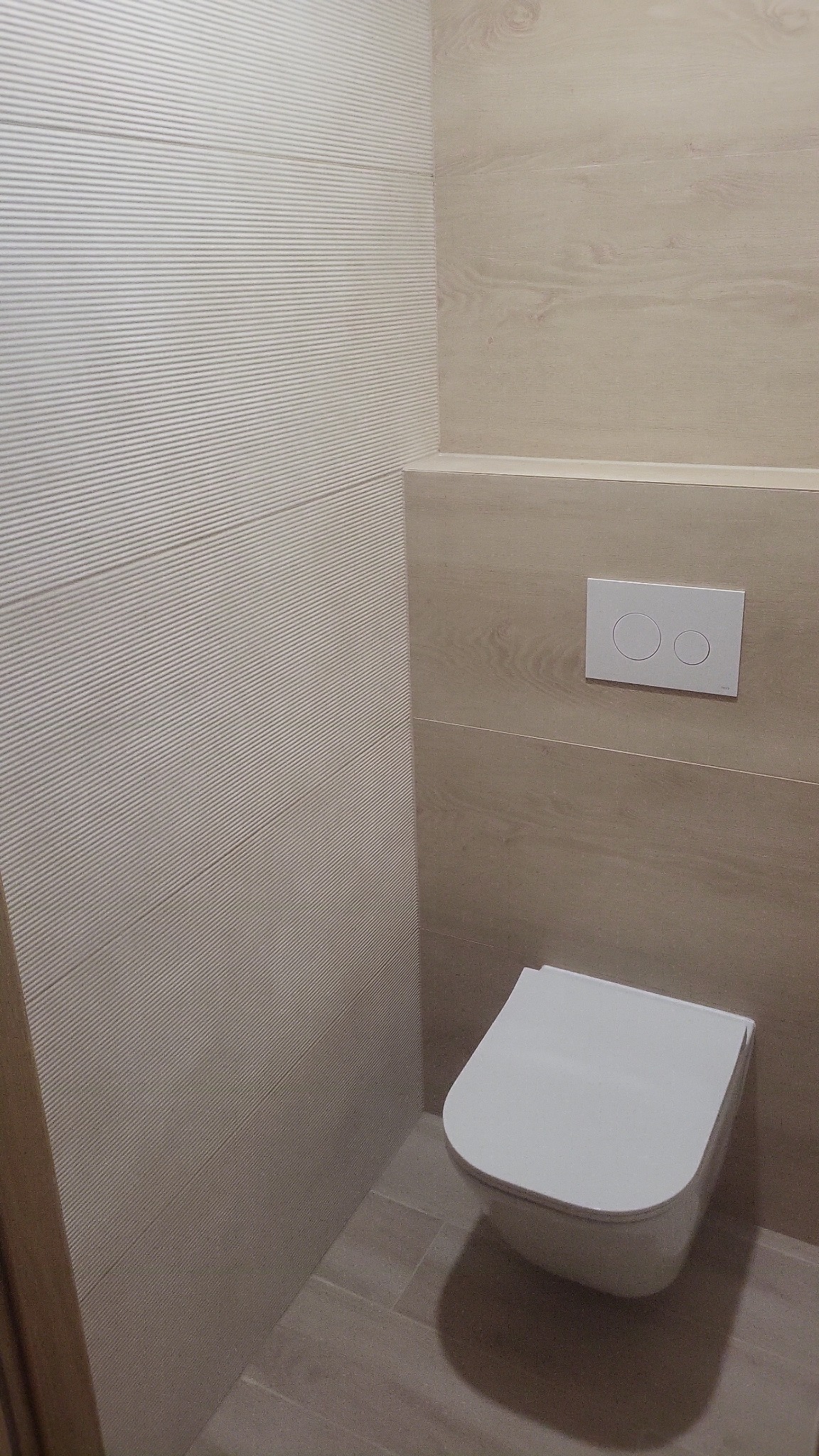 Nowoczesna toaleta podwieszana w jasnej łazience, z beżowymi płytkami imitującymi drewno i strukturalną ścianą. Minimalistyczny design i czysta estetyka.