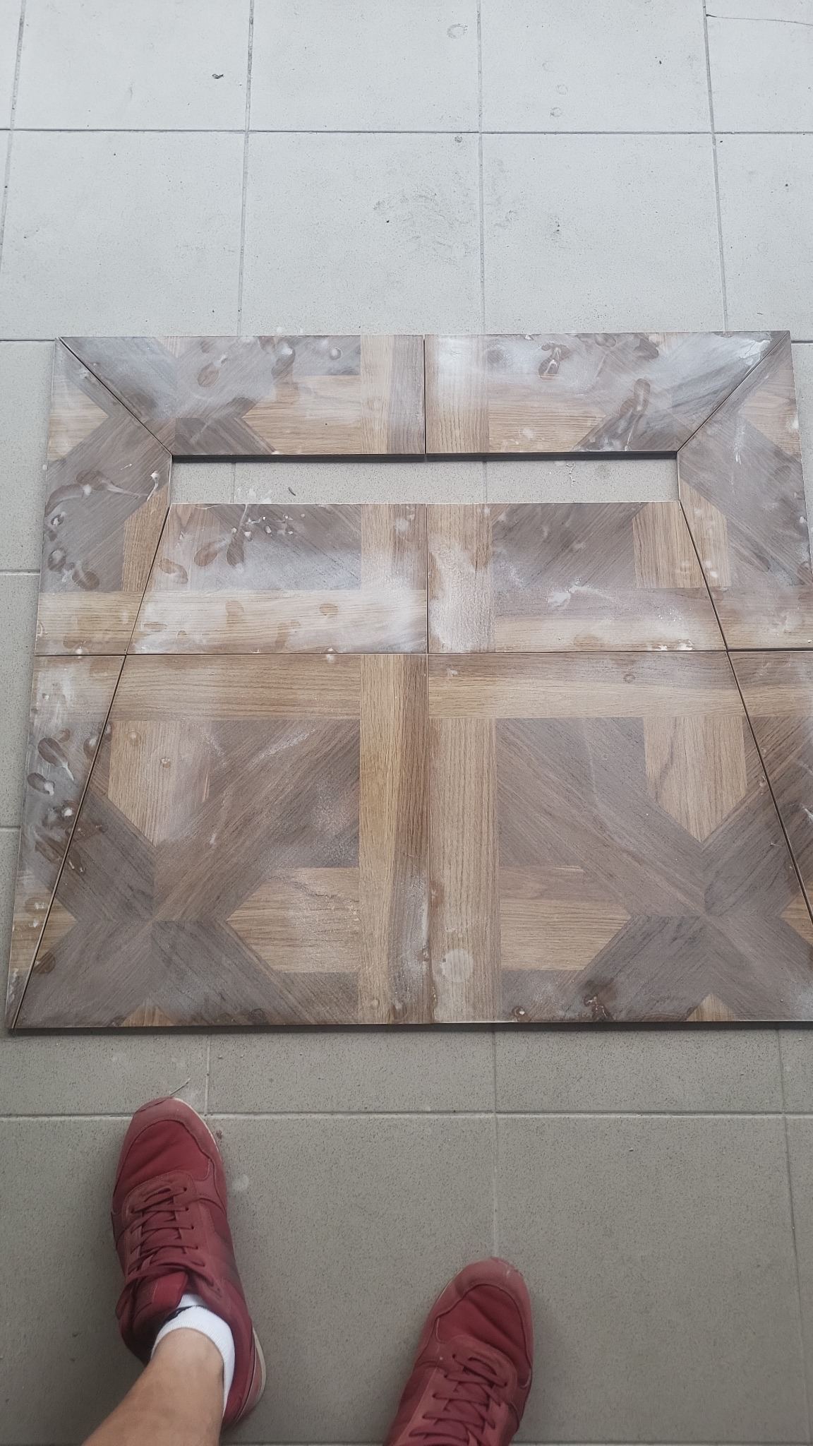 Ułożone płytki imitujące drewno w geometryczny wzór z widocznymi śladami zaprawy, na podłodze z szarych płytek. Widoczne buty osoby fotografującej.
