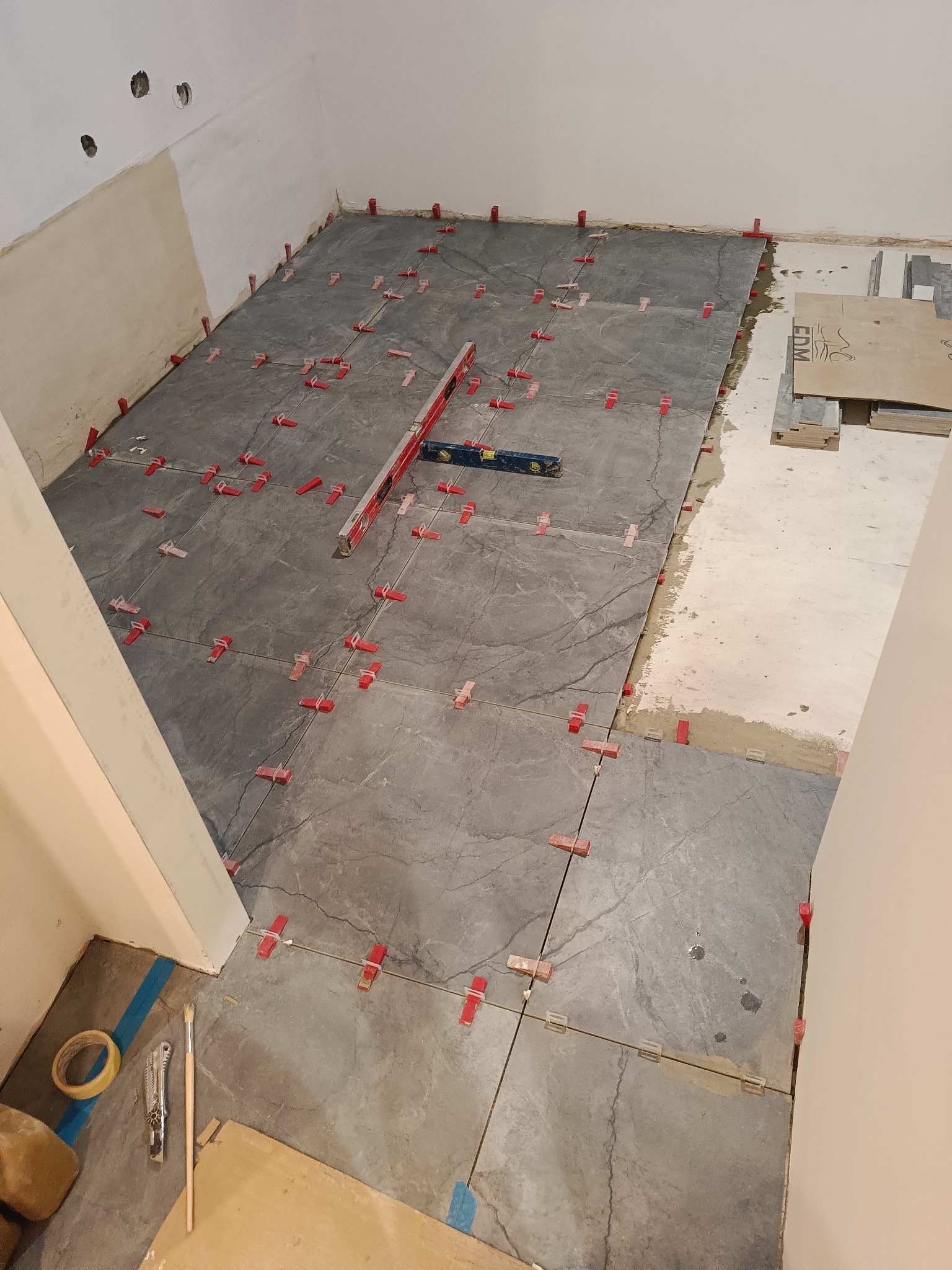 Układanie szarych płytek podłogowych z użyciem poziomicy i czerwonych klipsów poziomujących w trakcie prac wykończeniowych w pomieszczeniu.