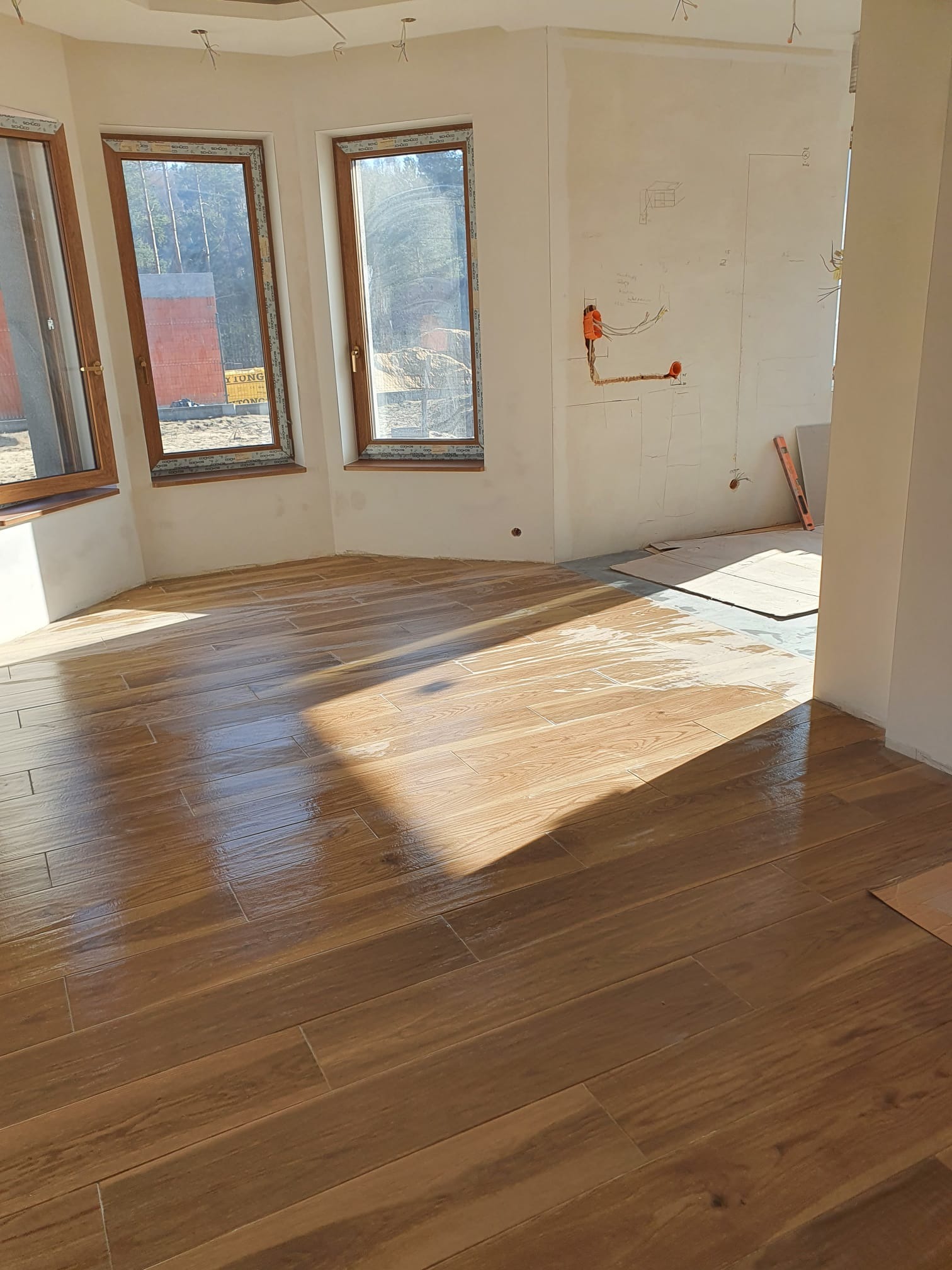 Ułożona podłoga z płytek imitujących drewno w jasnym pomieszczeniu z oknami. Ściany w trakcie wykończenia, widoczne instalacje elektryczne.