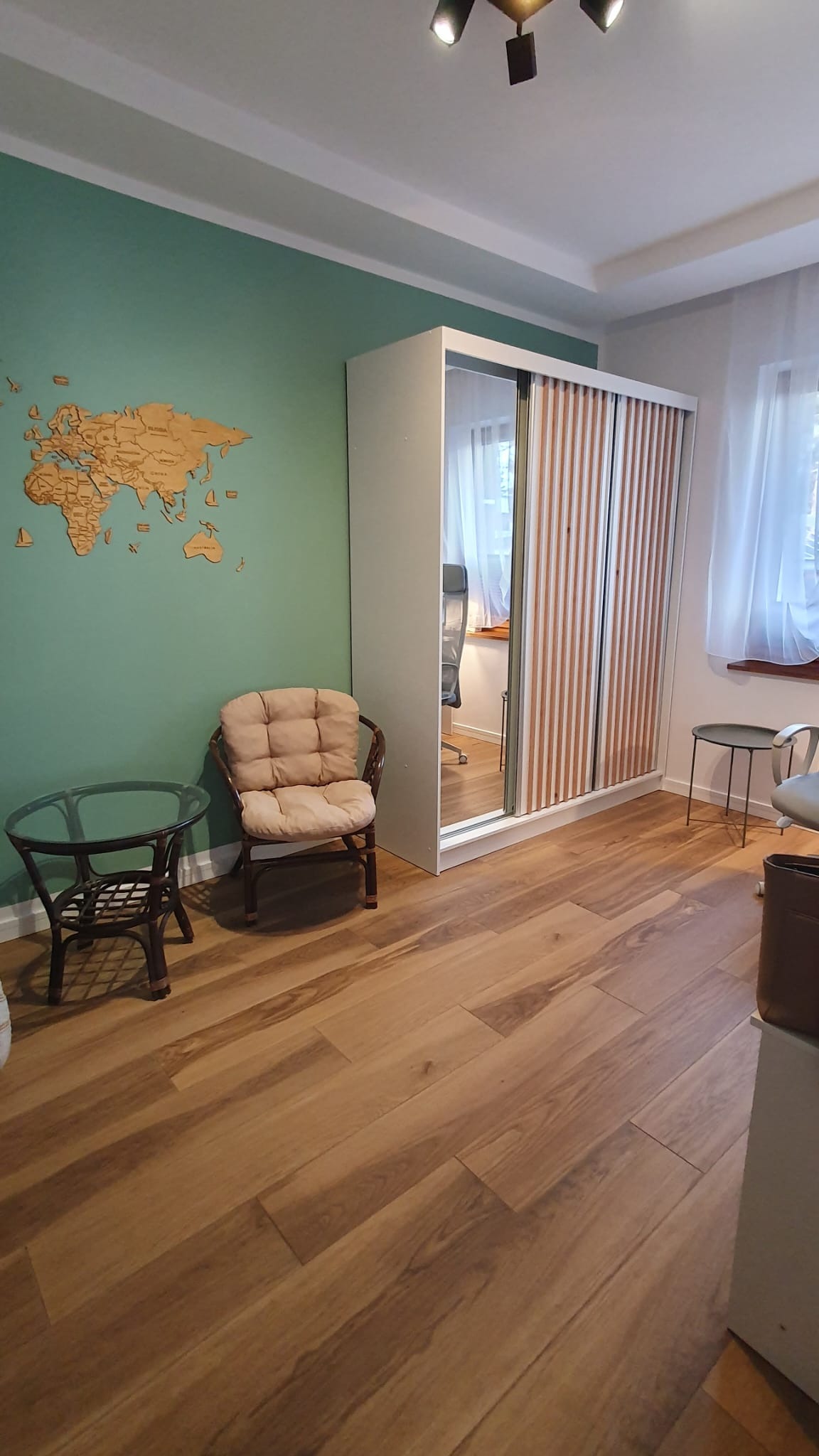 Pokój z drewnianą podłogą, szafą z lustrzanymi drzwiami przesuwnymi i drewnianą mapą świata na zielonej ścianie. W tle okno z białymi zasłonami.