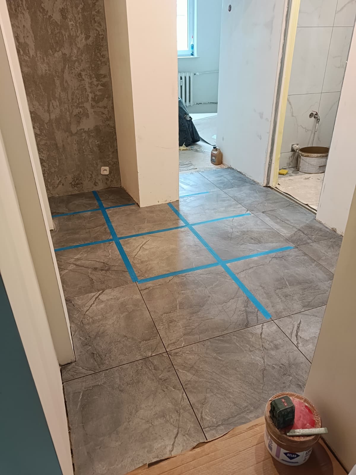 Szare płytki podłogowe z niebieską taśmą wyznaczającą wzór układania, widok na remontowane pomieszczenia i narzędzia glazurnicze, etap prac wykończeniowych.
