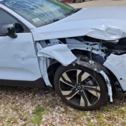 Powypadkowy, biały samochód marki Volvo z mocno uszkodzonym przodem, zwłaszcza błotnikiem i zderzakiem, stoi na żwirowym podłożu. Widoczne uszkodzenia mechaniczne.