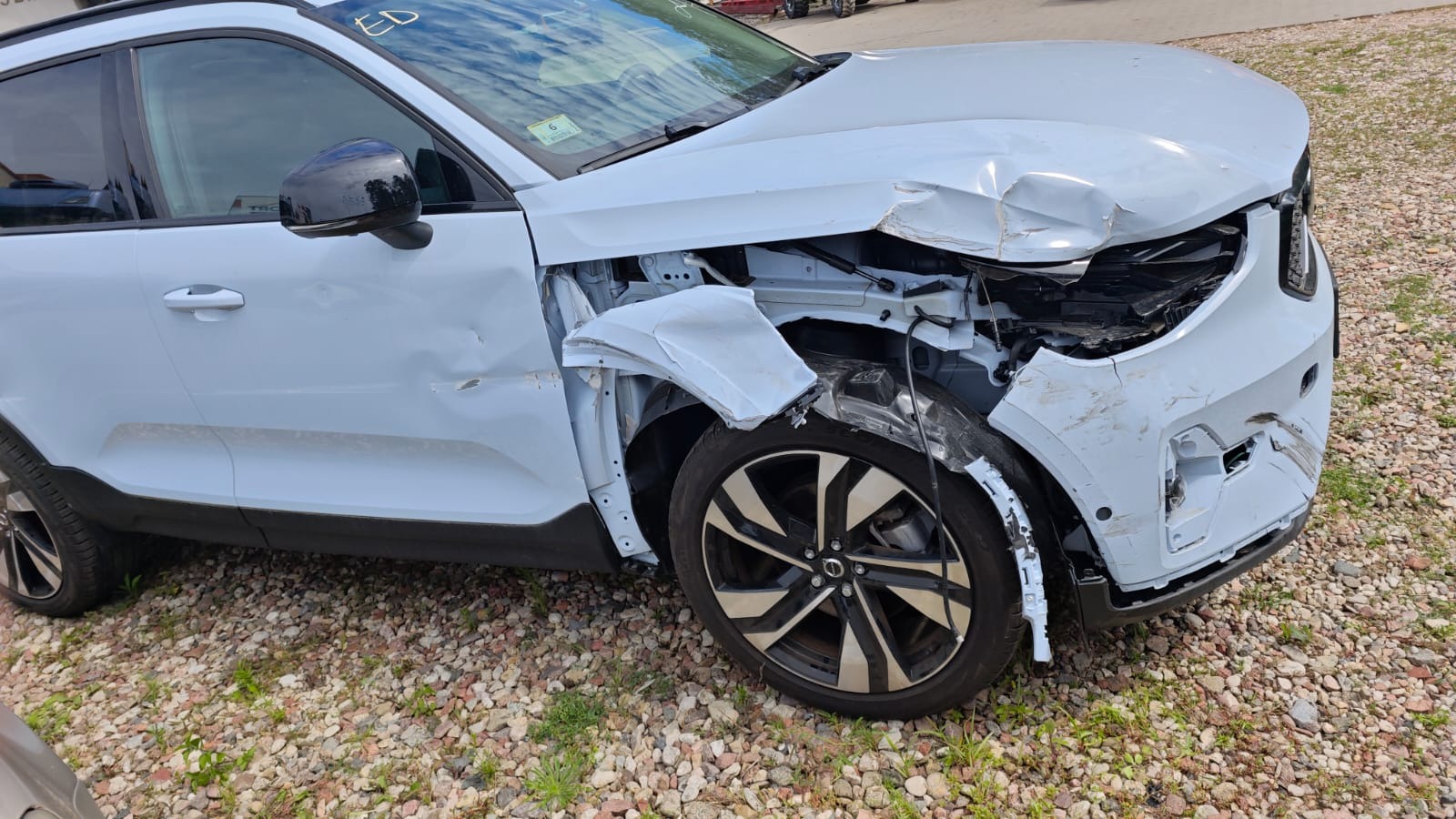 Powypadkowy, biały samochód marki Volvo z mocno uszkodzonym przodem, zwłaszcza błotnikiem i zderzakiem, stoi na żwirowym podłożu. Widoczne uszkodzenia mechaniczne.