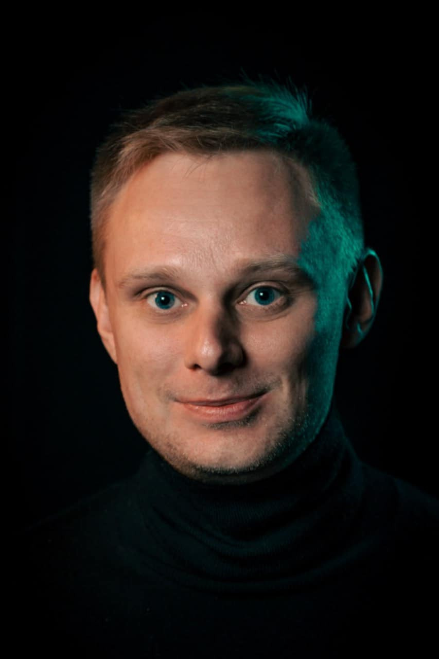 Portret mężczyzny z krótkimi blond włosami i niebieskimi oczami, ubranego w czarny golf, oświetlonego zielonkawym światłem z boku na ciemnym tle.