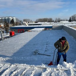 las bar Piotr Burda - Pracownik w trakcie odśnieżania płaskiego dachu budynku komercyjnego, używając łopaty i specjalnego narzędzia do usuwania śniegu. W tle widoczne silosy i zabudowania.