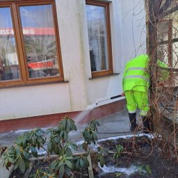 las bar Piotr Burda - Pracownik w jaskrawozielonym kombinezonie czyści elewację domu myjką ciśnieniową. Widoczne zabrudzenia na ścianie i woda spływająca po elewacji.