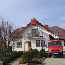 las bar Piotr Burda - Pracownik w czerwonym podnośniku koszowym przy białym domu z brązowym dachem. Widoczny numer telefonu na ramieniu podnośnika. Błękitne niebo w tle.