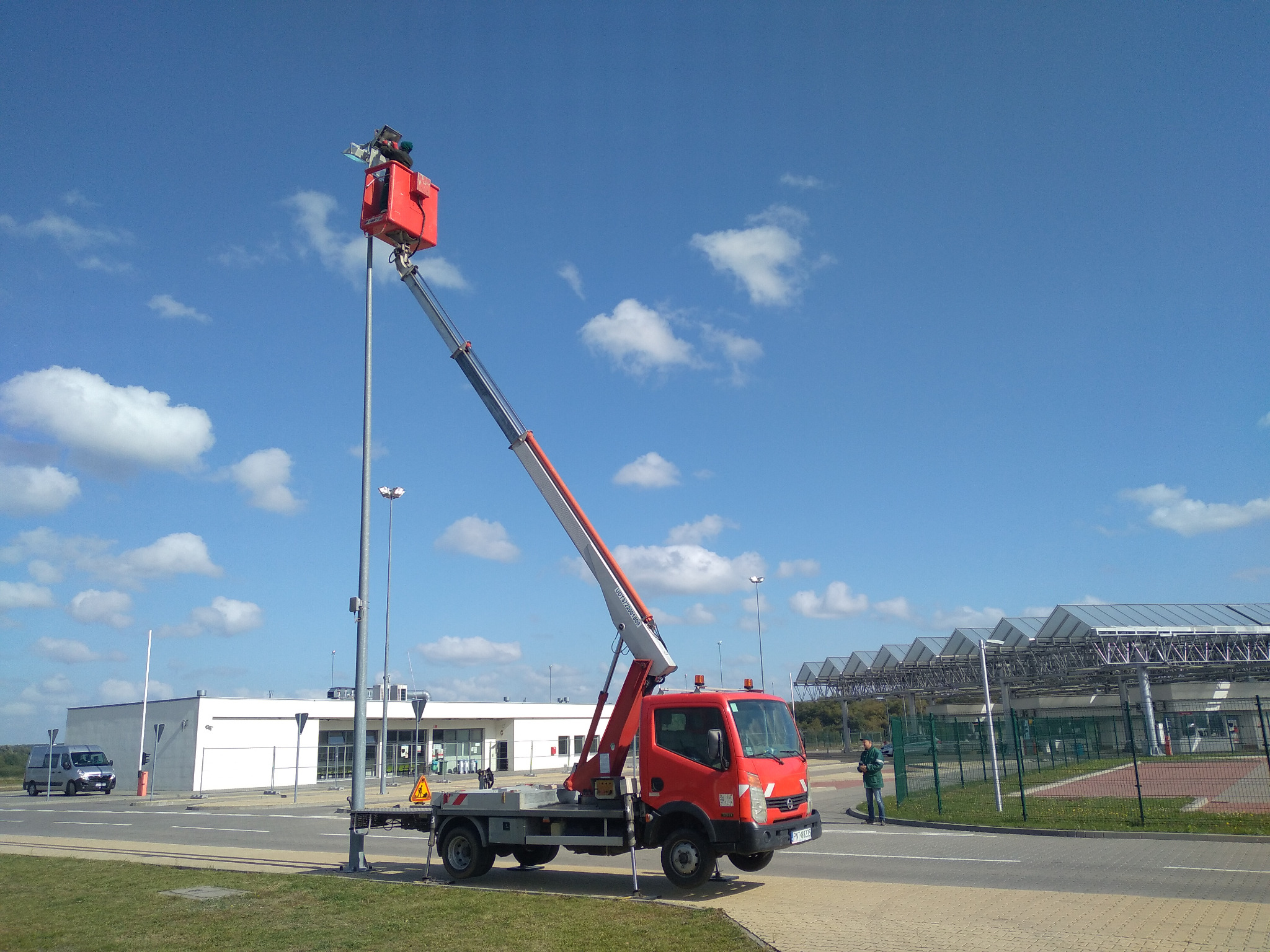 Czerwony podnośnik koszowy na tle błękitnego nieba z chmurami, pracownik w koszu przy słupie oświetleniowym, budynek w tle. Dzień roboczy, widok z dołu.