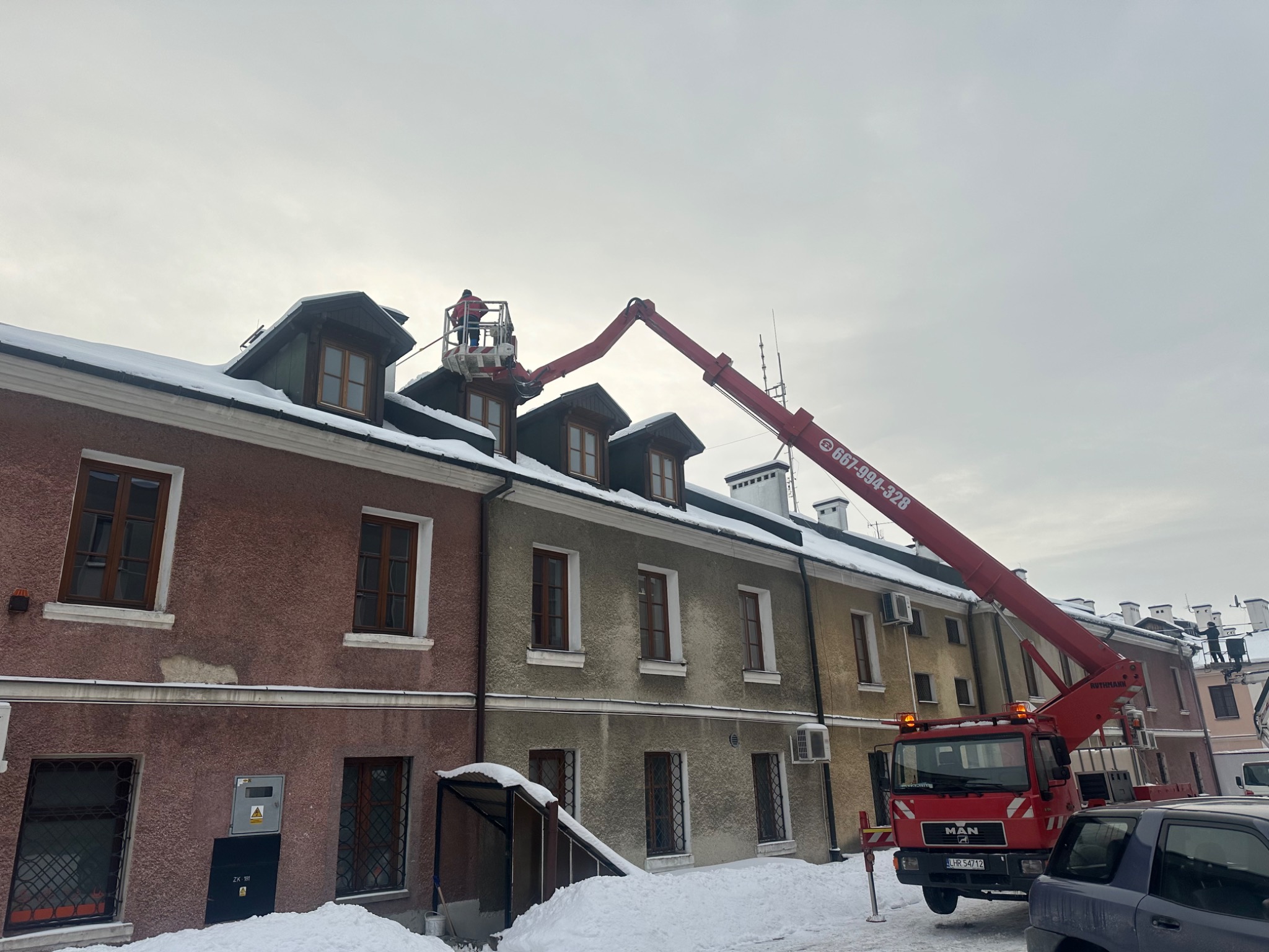 Czerwony podnośnik koszowy MAN na tle budynku z zaśnieżonym dachem, z pracownikiem w koszu na wysokości. Zimowy dzień, pochmurne niebo.