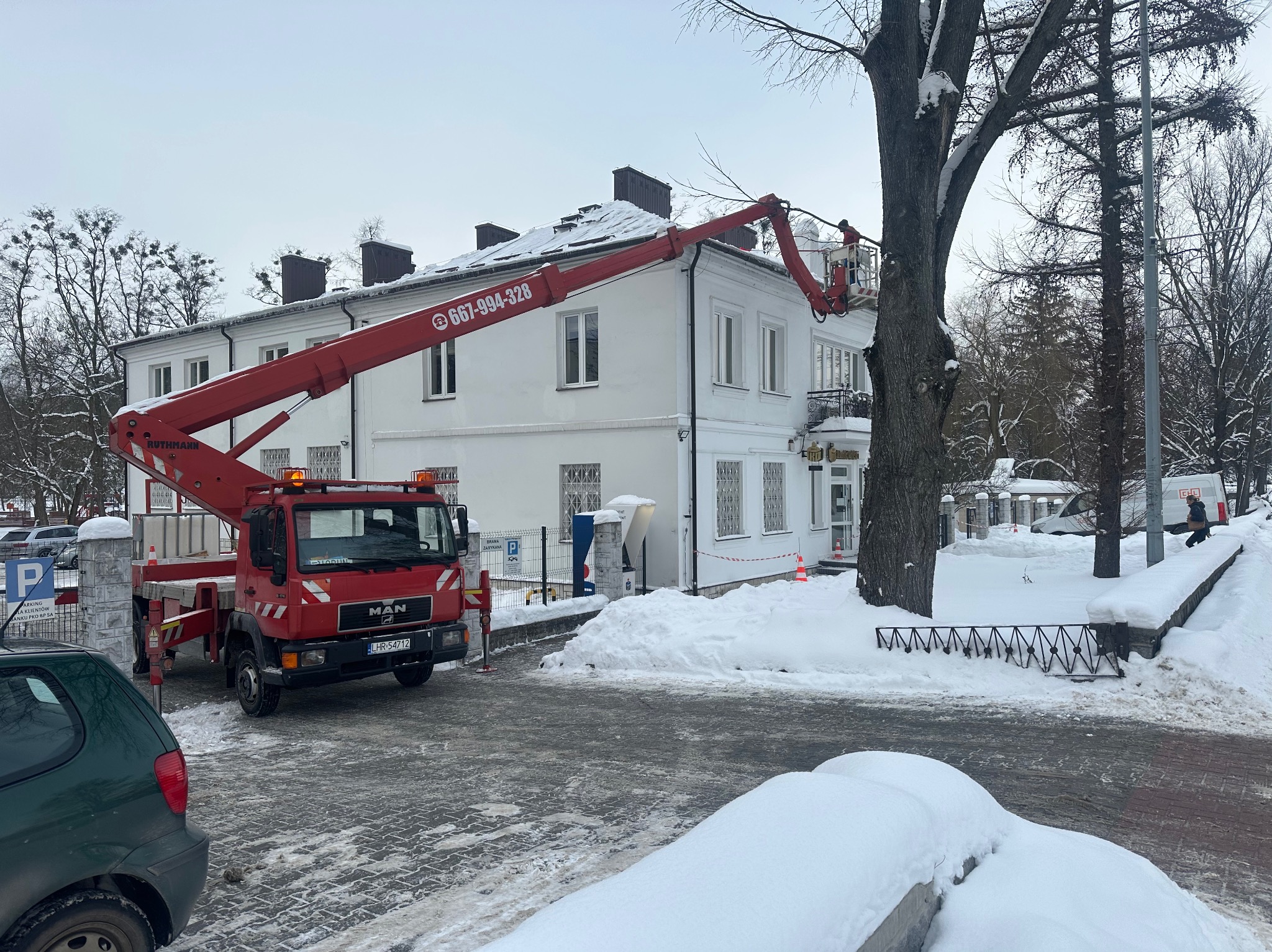 Czerwony podnośnik koszowy MAN na tle białego budynku w zimowej scenerii. Pracownik w koszu przycina gałęzie drzewa. Widoczny numer telefonu na ramieniu podnośnika.
