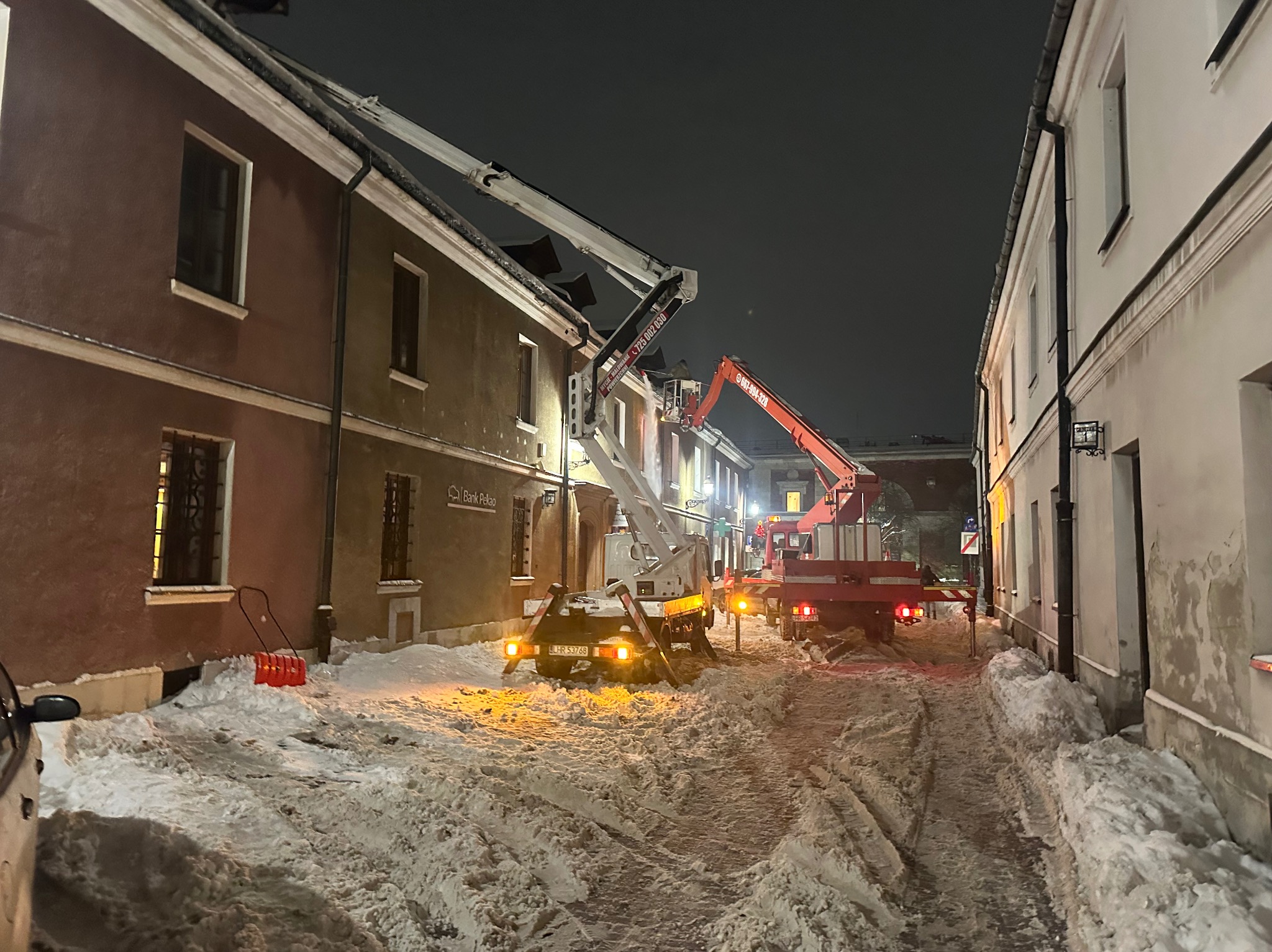 Dwa podnośniki koszowe oświetlają budynki podczas odśnieżania dachów w zimowej scenerii miejskiej nocą. Śnieg zalega na ulicy, a w tle widoczne są światła miasta.
