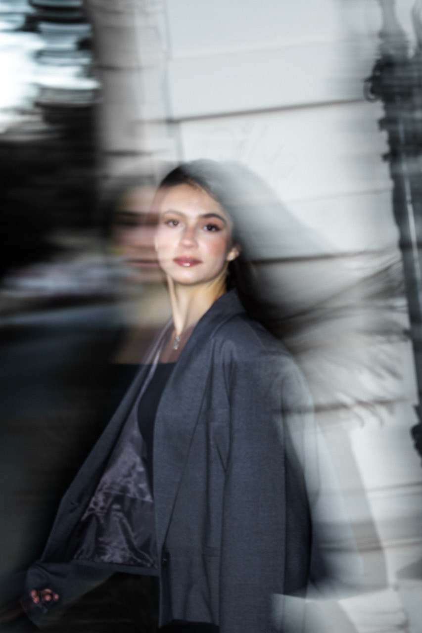 Portret młodej kobiety w szarym żakiecie, uchwycony w ruchu z efektem rozmycia. Ujęcie z dołu, jasne tło z elementami architektonicznymi.