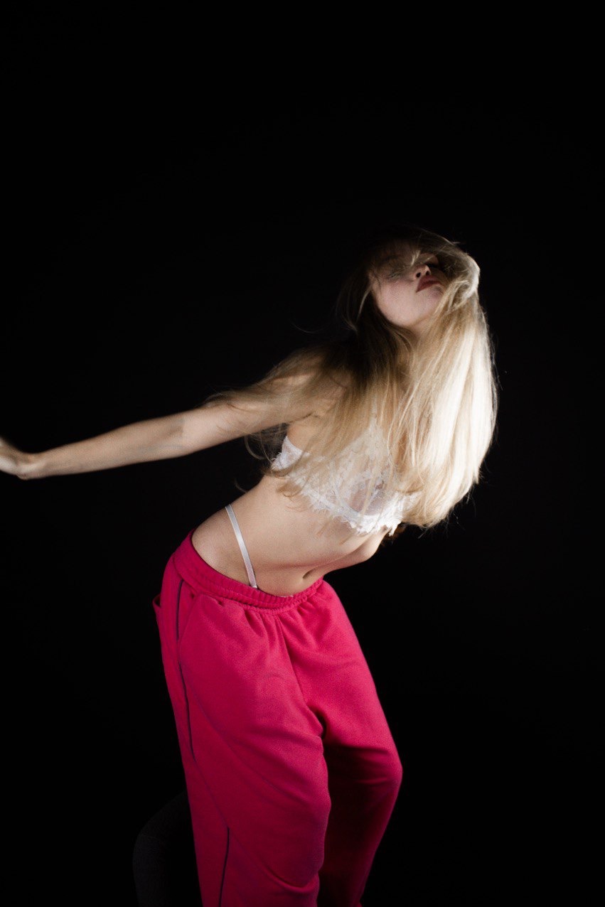 Dynamiczne ujęcie kobiety z rozwianymi blond włosami, w białym koronkowym staniku i różowych spodniach dresowych na czarnym tle, uchwycone w ruchu podczas sesji.
