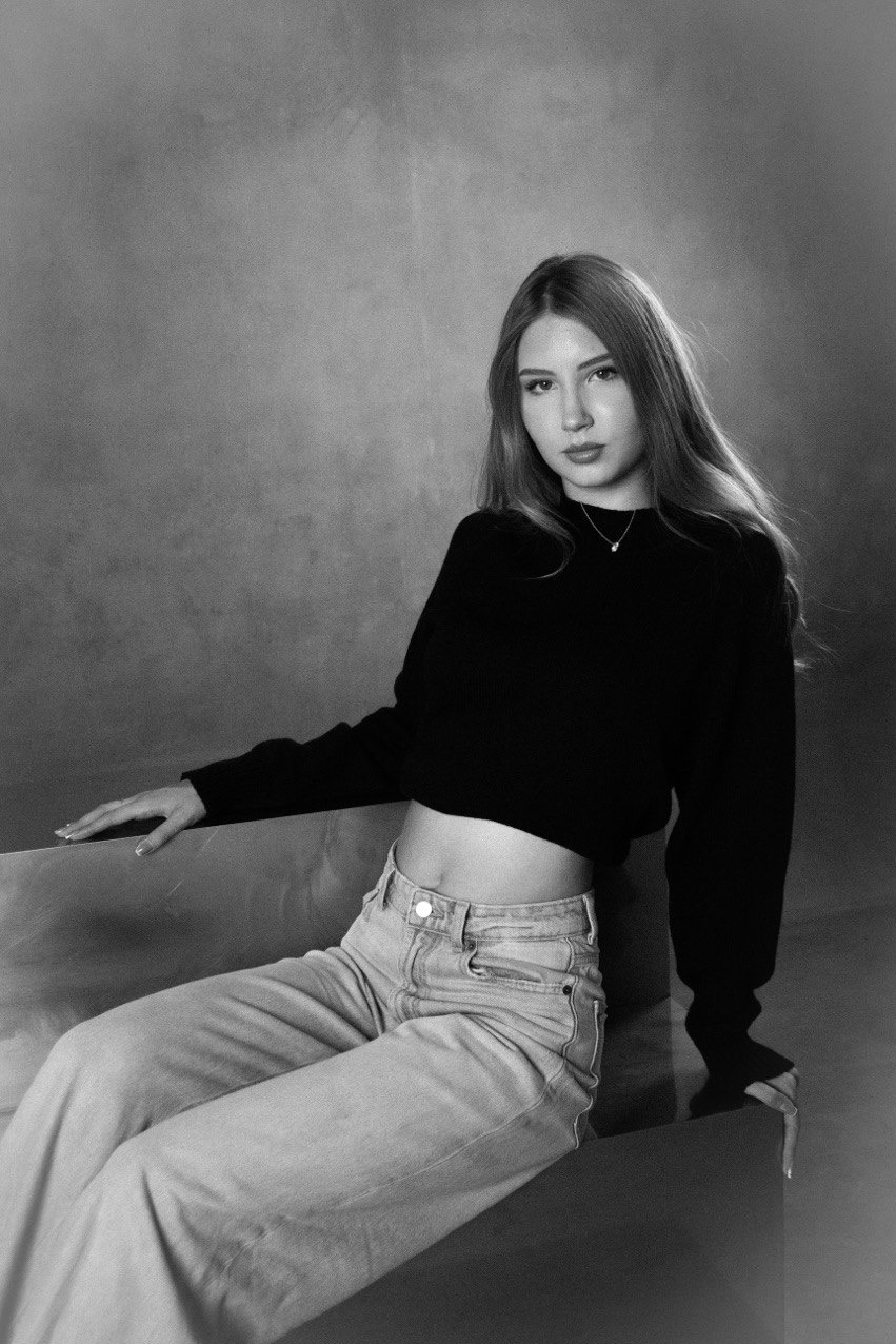 Portret młodej kobiety w czarno-białej sesji. Modelka siedzi w jeansach i czarnym swetrze, z delikatnym uśmiechem. Stonowane tło podkreśla naturalność i subtelność zdjęcia.