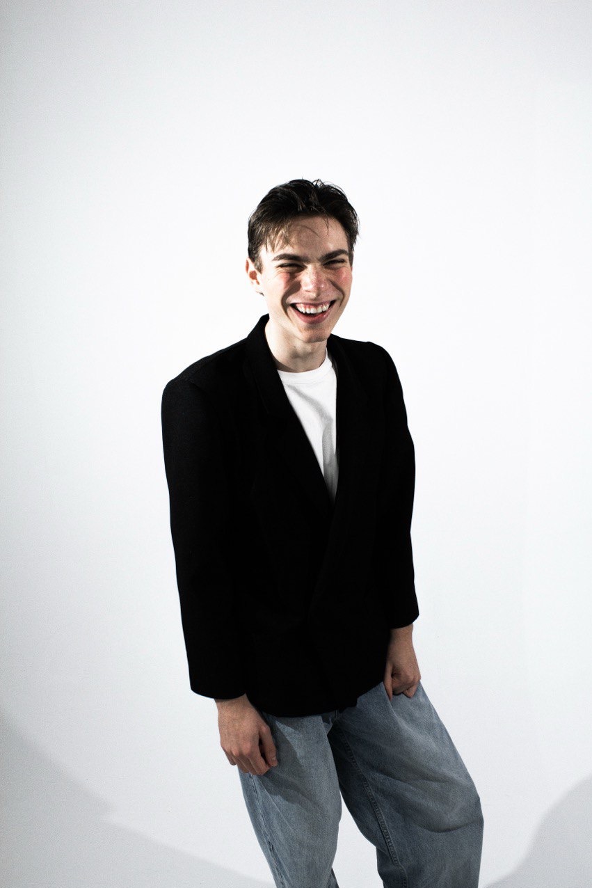 Uśmiechnięty młody mężczyzna w czarnej marynarce i jeansach na białym tle. Zdjęcie studyjne z widocznymi piegami i radosnym wyrazem twarzy.