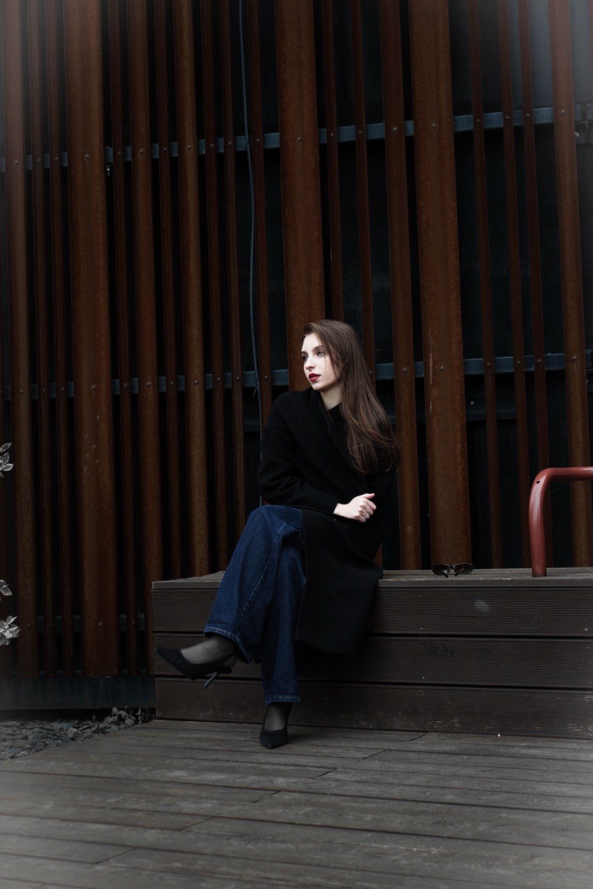 Zamyślona brunetka w czarnym płaszczu i dżinsach siedzi na ławce na tle rustykalnej ściany. Ciemna tonacja, stonowane kolory, elegancki, miejski styl.