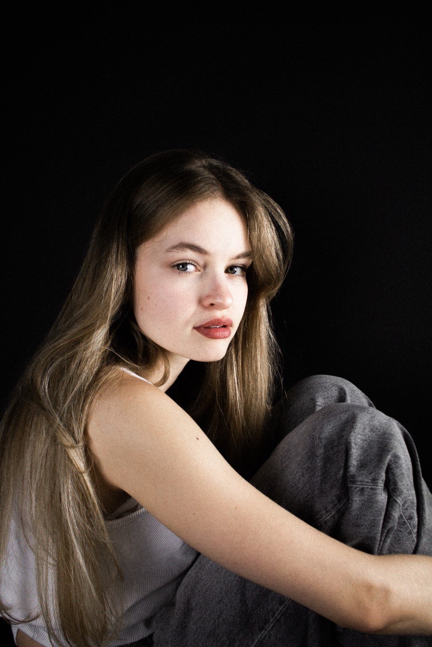 Portret młodej kobiety z długimi, jasnymi włosami na czarnym tle. Ubrana w biały top i szare jeansy, obejmuje kolana. Delikatny makijaż, naturalne światło.