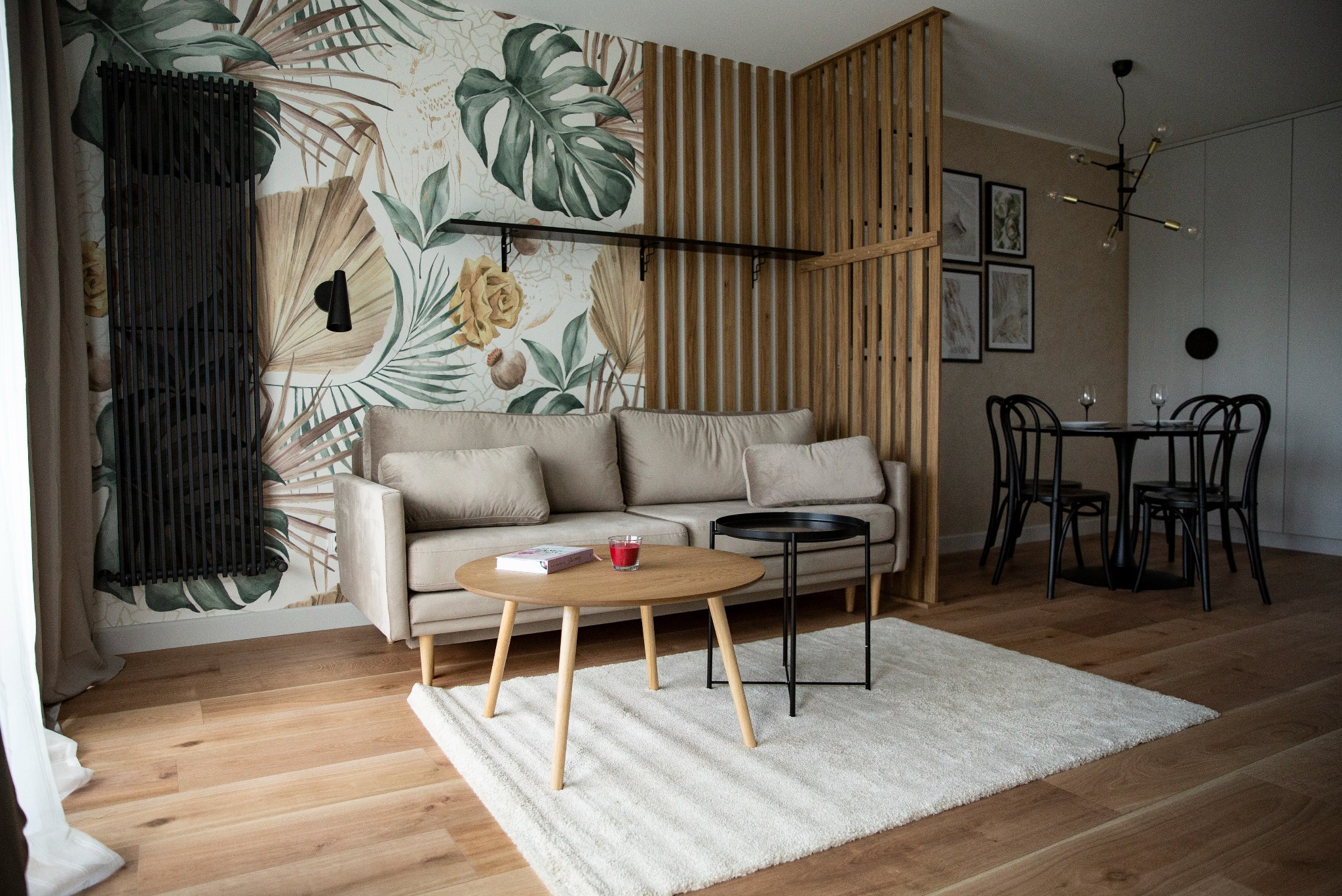 Salon z beżową sofą, okrągłymi stolikami i ścianą w tropikalne wzory. Czarny grzejnik i drewniana ażurowa ścianka działowa dodają wnętrzu charakteru.