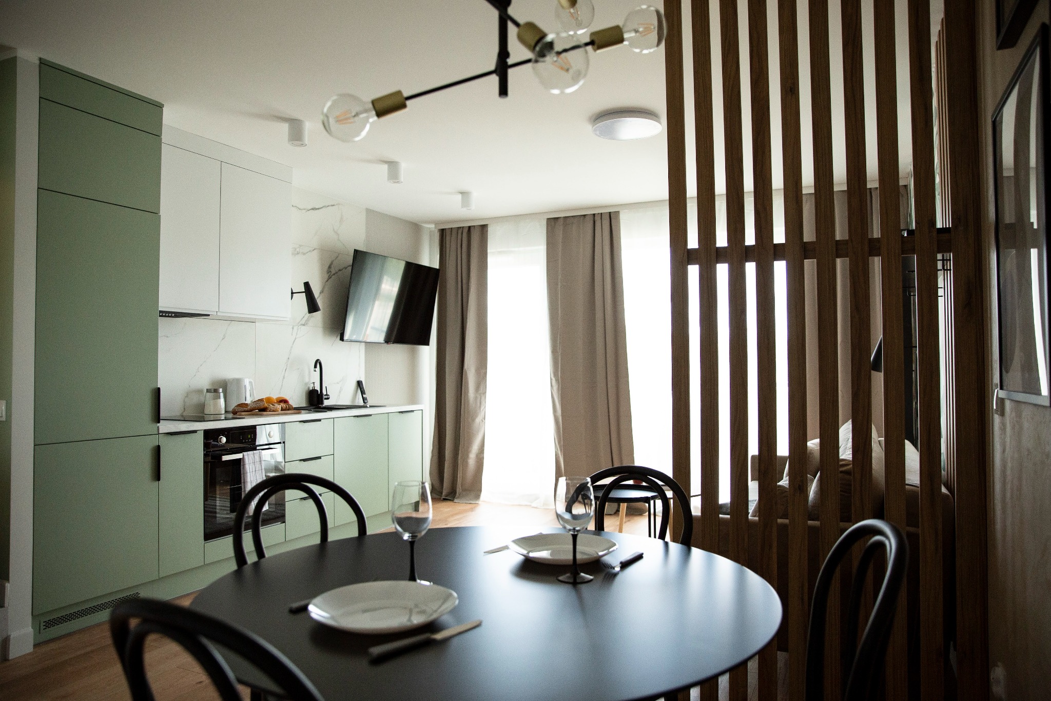 Nowoczesna kuchnia z zielonymi szafkami i marmurowym blatem, widok na okrągły stół z nakryciem i drewnianą ażurową ścianką działową w tle.