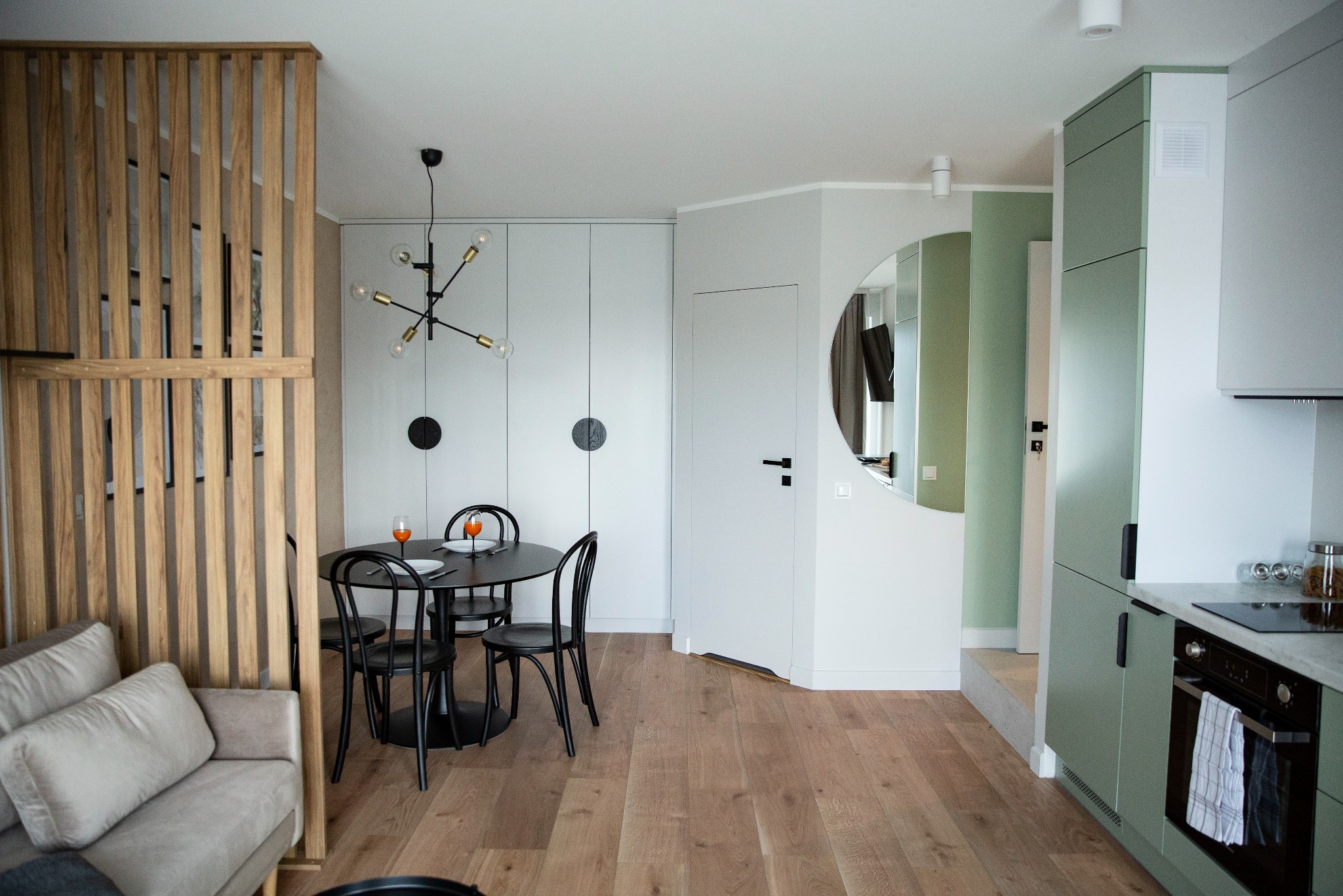 Jasne wnętrze mieszkania z drewnianą podłogą i ścianami w odcieniach szarości i zieleni. Widoczny stół z krzesłami, sofa, kuchnia i okrągłe lustro.