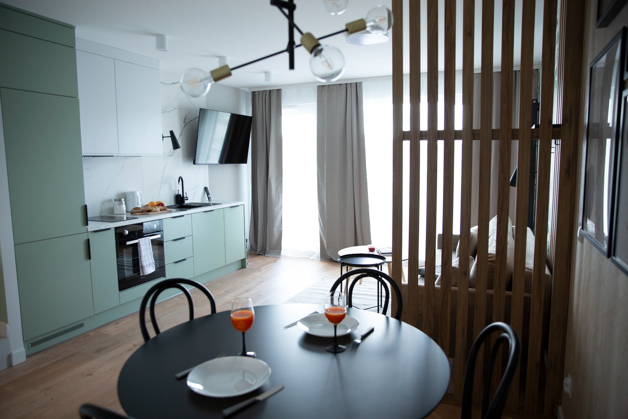 Nowoczesna kuchnia z salonem: pastelowe szafki, czarny stół z nakryciem, drewniana ścianka działowa i widok na jasny pokój dzienny. Designerski wystrój wnętrza.