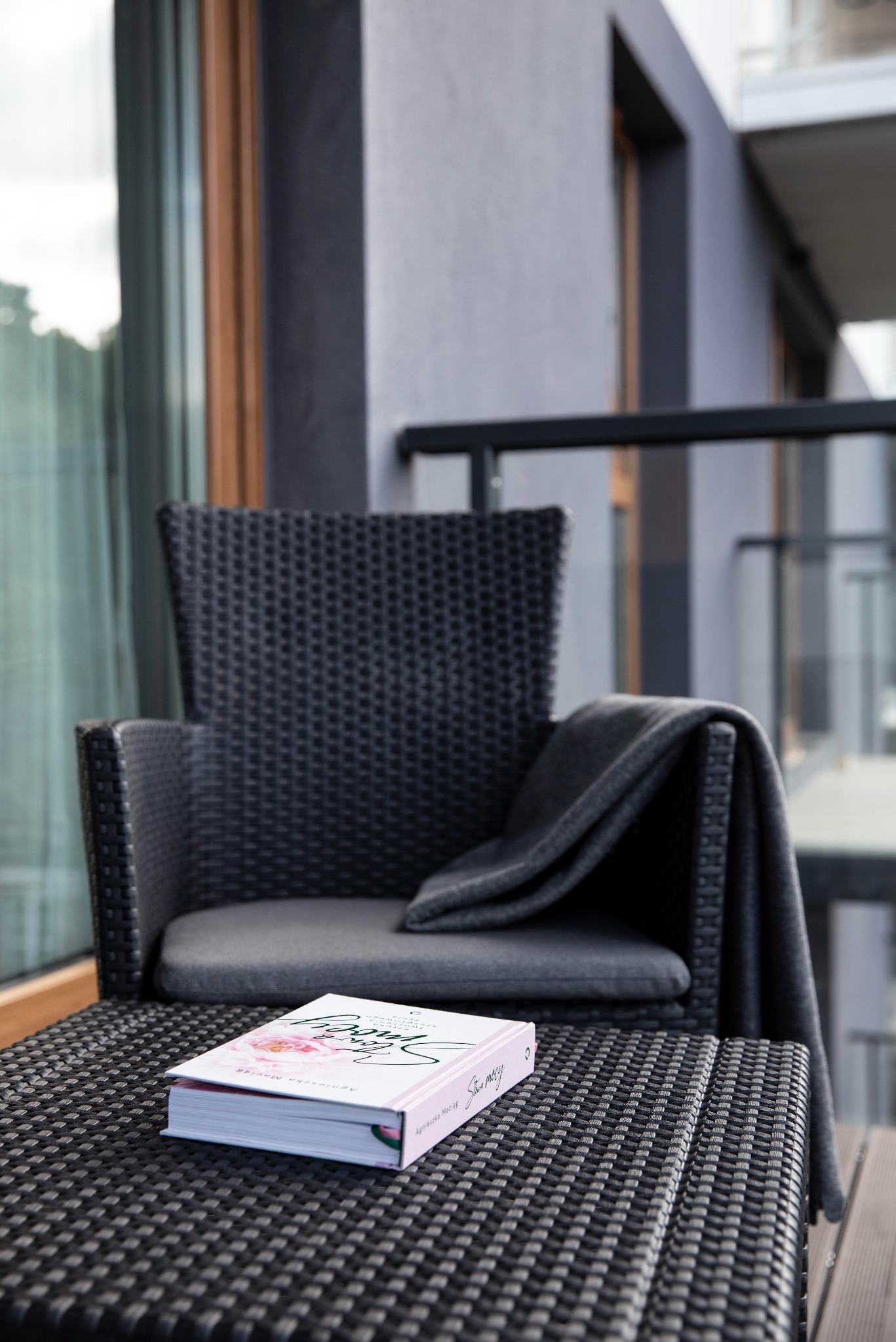 Stylowy balkon z krzesłem z rattanu, pledem i książką na stoliku. Nowoczesne wykończenie balkonu z szarymi ścianami i szklaną balustradą.
