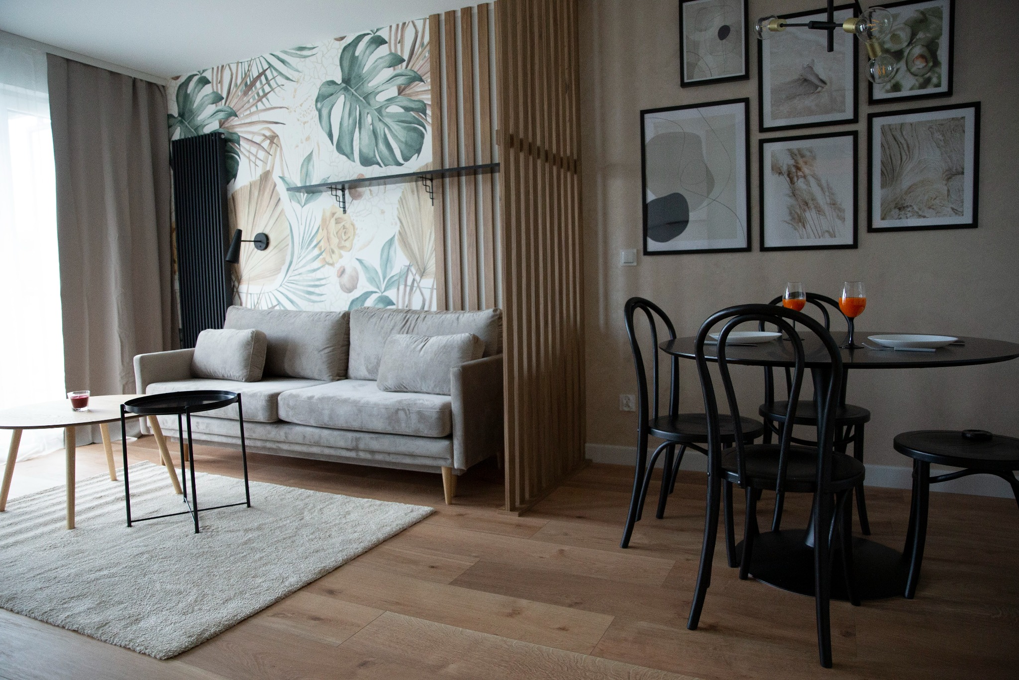 Jasne wnętrze salonu z sofą, stolikiem kawowym na dywanie i okrągłym stołem z czarnymi krzesłami. Na ścianie dekoracyjna tapeta w liście i galeria obrazów.