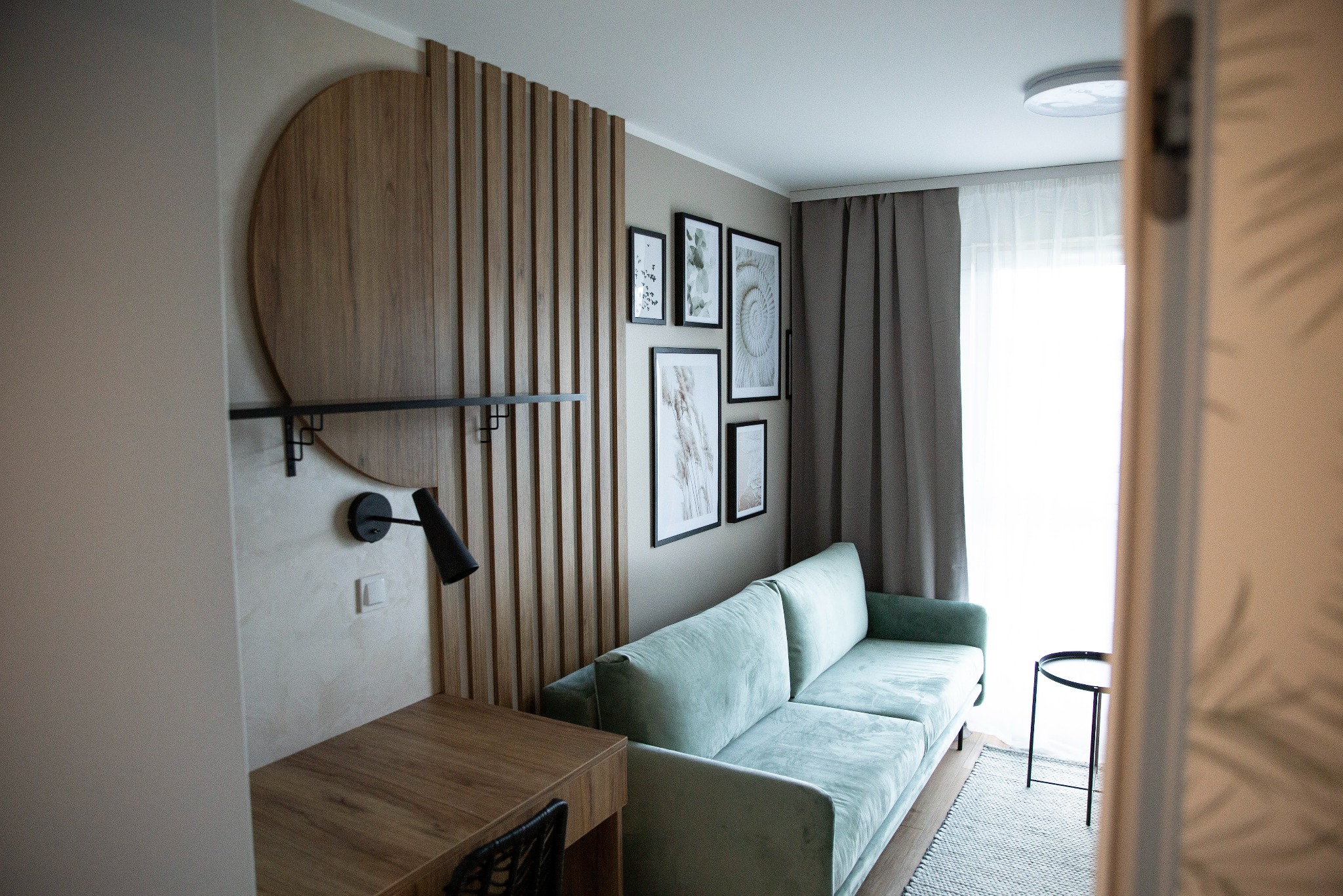 Stylowy pokój dzienny z zieloną sofą, drewnianą ścianką działową i dekoracjami ściennymi. Na biurku stoi lampka. Widok zza drzwi.